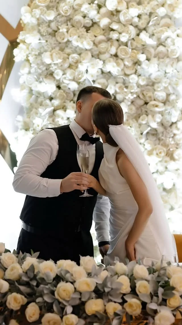 A bride and groom sharing a kiss at their wedding reception, with a large floral backdrop of white roses behind them and a table decorated with white roses in the foreground.