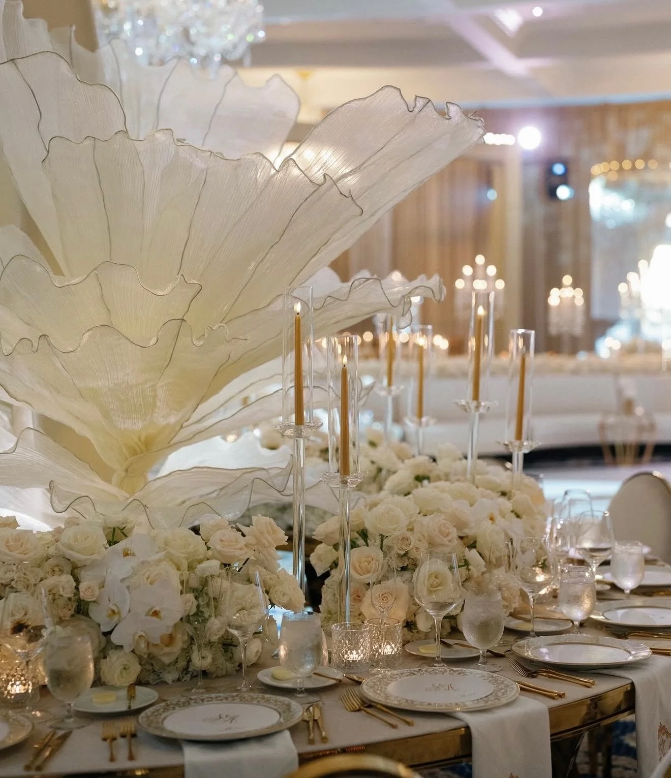 Elegant banquet table decorated with white flowers, tall candleholders with lit candles, and a large, decorative white leaf centerpiece in a softly lit setting.