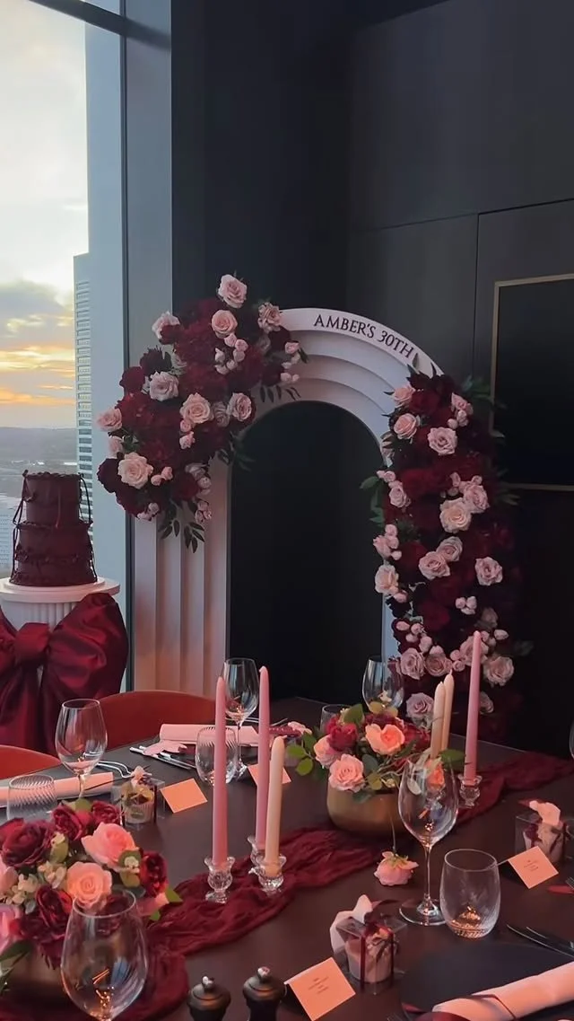 Decorated dining table with pink and red floral arrangements, pink candles, wine glasses, and small favors, set for a 30th birthday celebration named Amber, with a floral arch and a cake in the background.