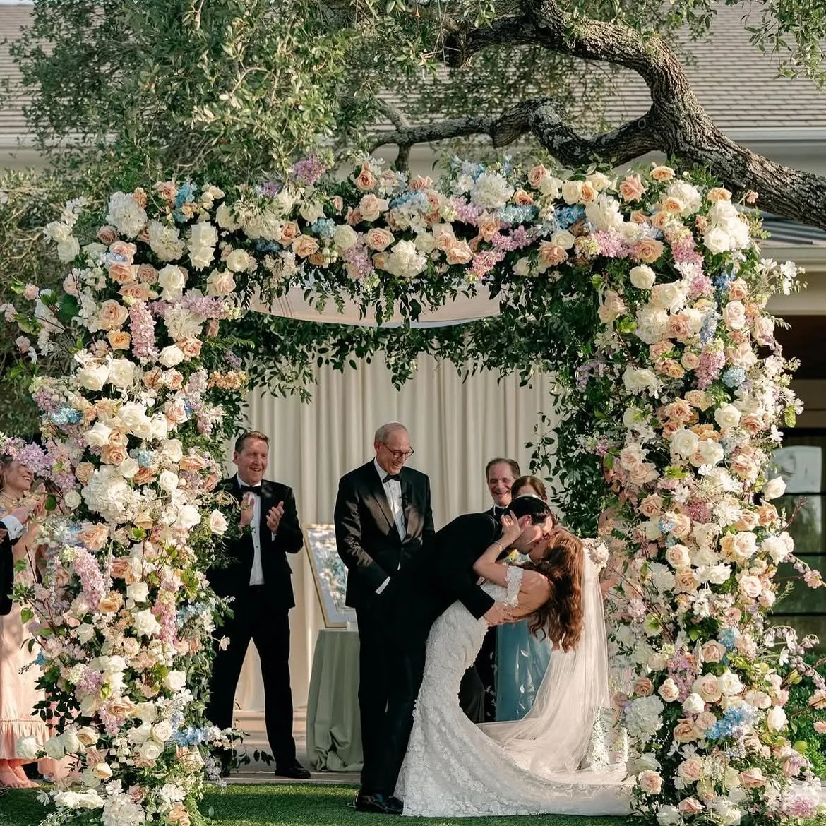 A wedding ceremony under a floral arch with a couple sharing a kiss, surrounded by guests clapping and smiling.