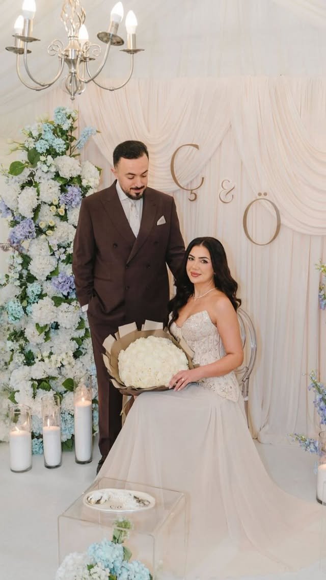 A couple celebrating a wedding with elegant floral decorations, candles, and a chandelier, with the bride seated holding a large bouquet of white flowers and the groom standing beside her.