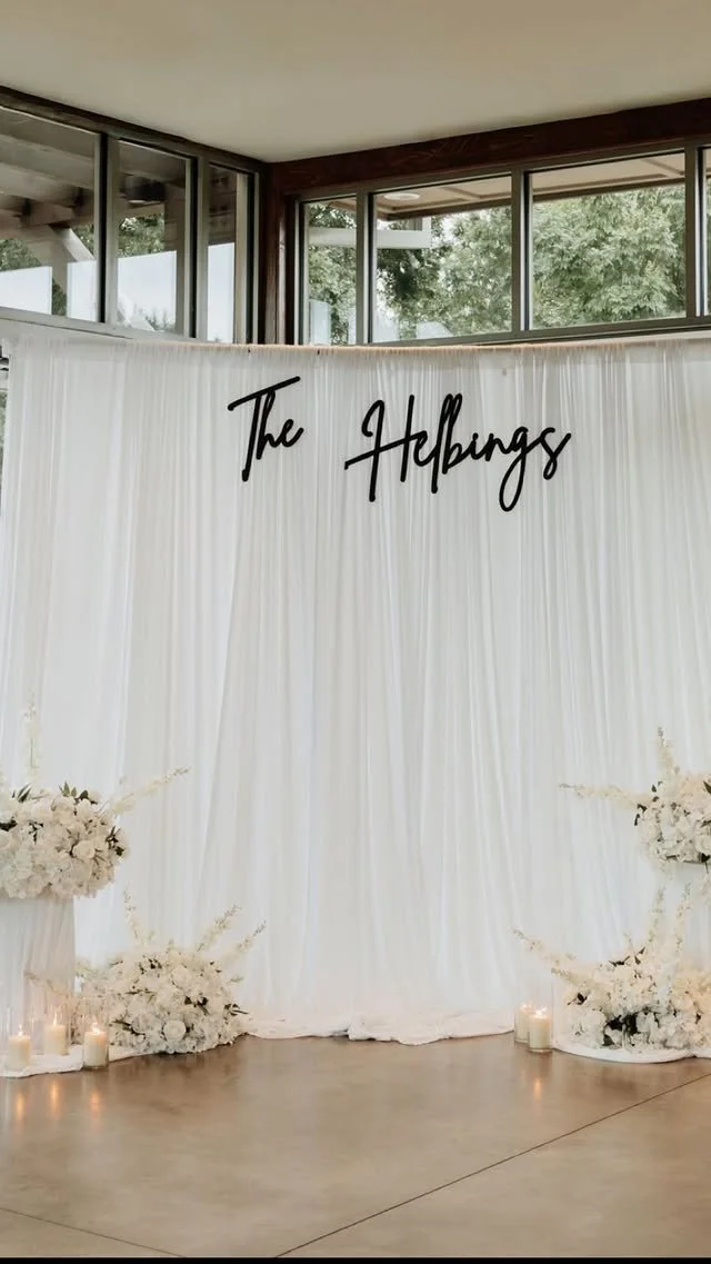 Wedding or event backdrop with white drapes, floral arrangements, and candles, featuring the text 'The Hebings' in black cursive.