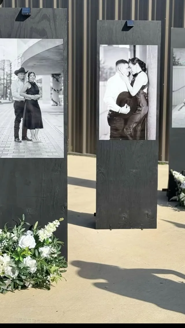 Outdoor photo display of black and white photographs on wooden panels, with white flowers at the base, at a memorial or event.