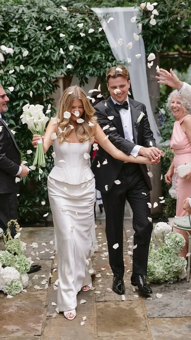 Wedding couple walking under falling flower petals, surrounded by happy guests, in an outdoor garden setting.