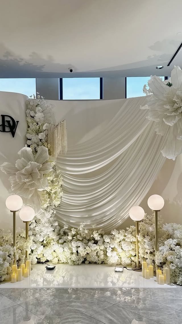Elegant wedding backdrop with white drapes, large white paper flowers, floral arrangements, and soft lighting.