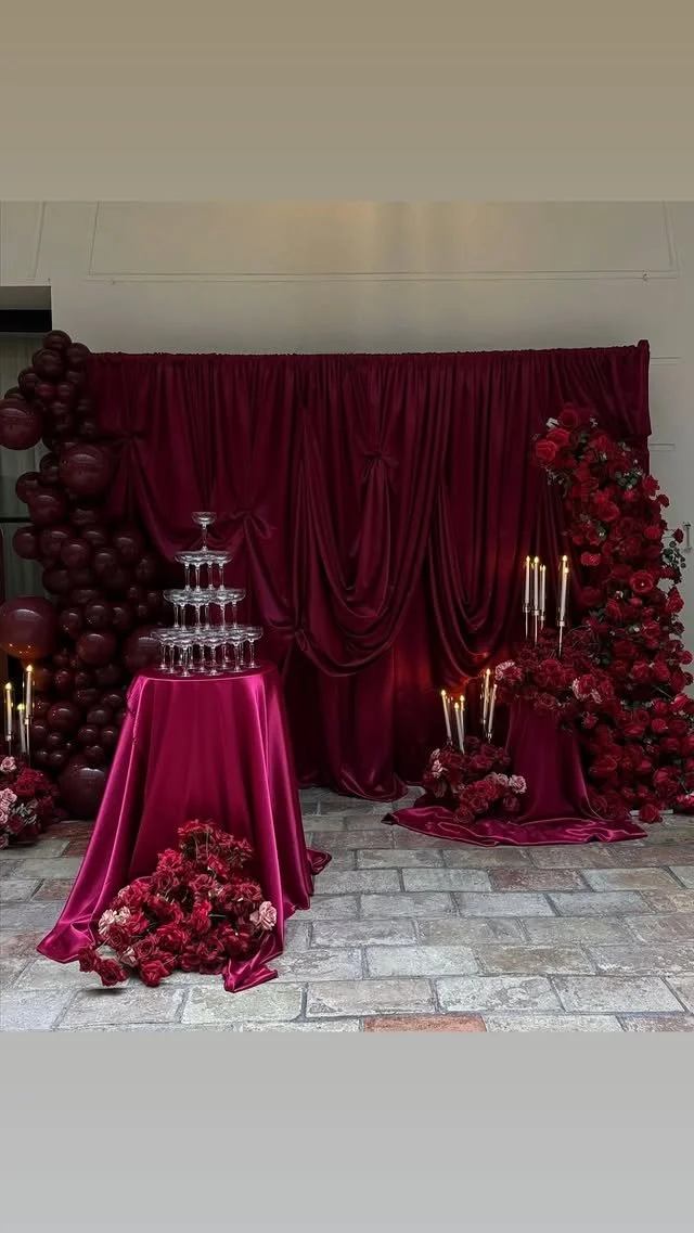 Decorative backdrop with rich burgundy curtains, balloons, and floral arrangements with candles for celebration or event.