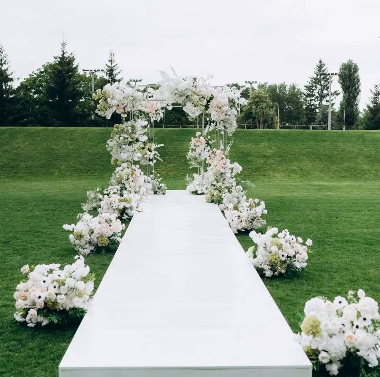 A white wedding aisle lined with floral arrangements leading to a flower arch on a green outdoor field.