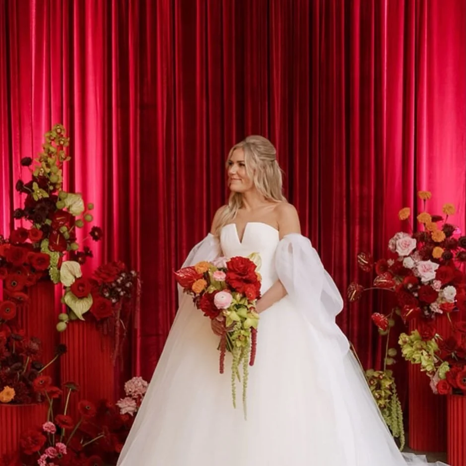 A bride in a white wedding gown holding a bouquet of red and pink flowers, standing in front of a red curtain backdrop decorated with various colorful floral arrangements.