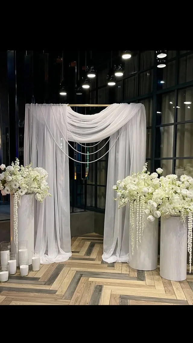 Elegant wedding arch with sheer white drapes and floral arrangements on large white vases, complemented by candles on the wooden floor.