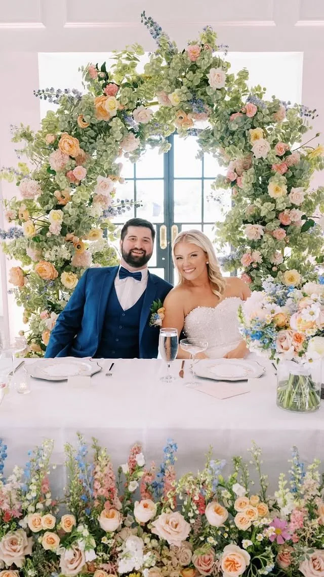 A bride and groom sitting at a decorated wedding table with a large floral arch behind them, smiling at the camera.