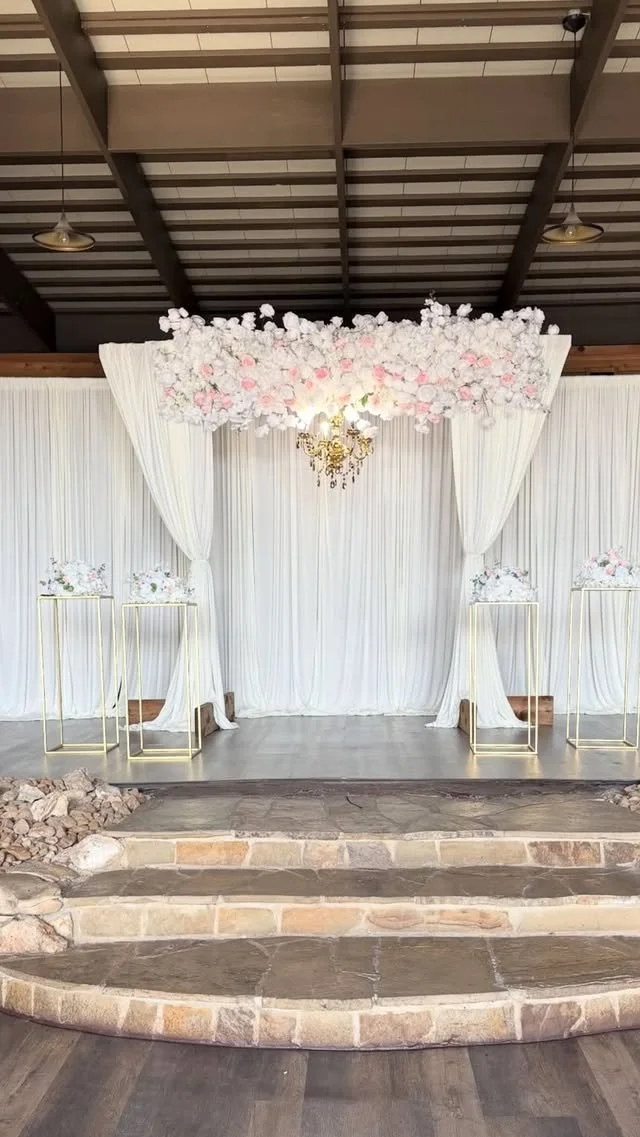 Wedding altar with white drapery, floral arrangement overhead, gold chandeliers, and decorative tables with floral arrangements, set on stone steps with a rustic ceiling.