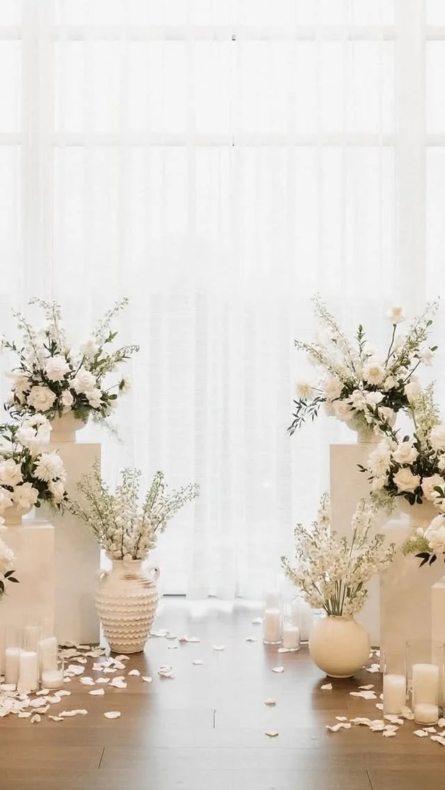 Elegant floral arrangement with white flowers and greenery in white vases, candles, and rose petals scattered on the floor near a white curtain backdrop.