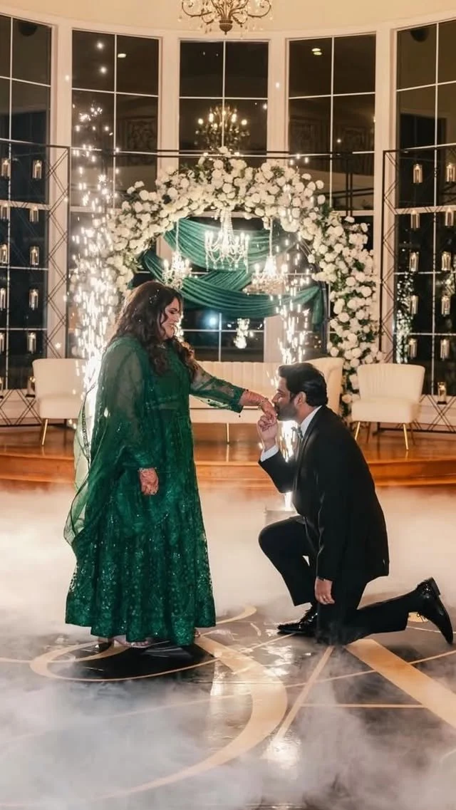 A couple at a wedding or celebration, with the man kneeling and holding the woman's hand, surrounded by sparks and fireworks, in a decorated indoor venue decorated with flowers and chandeliers.