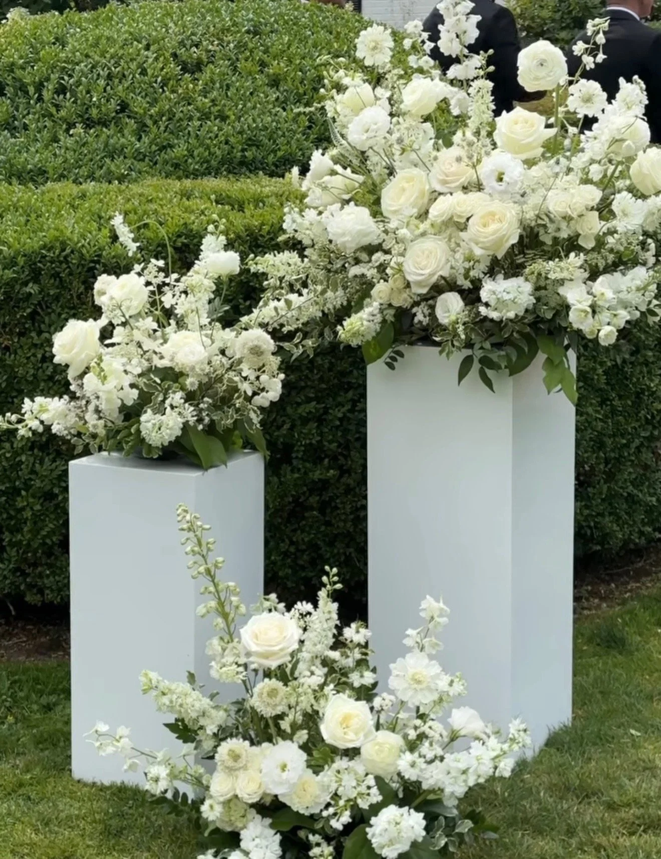 White floral arrangements on tall white pedestals, with a background of green bushes and a formal event setting.