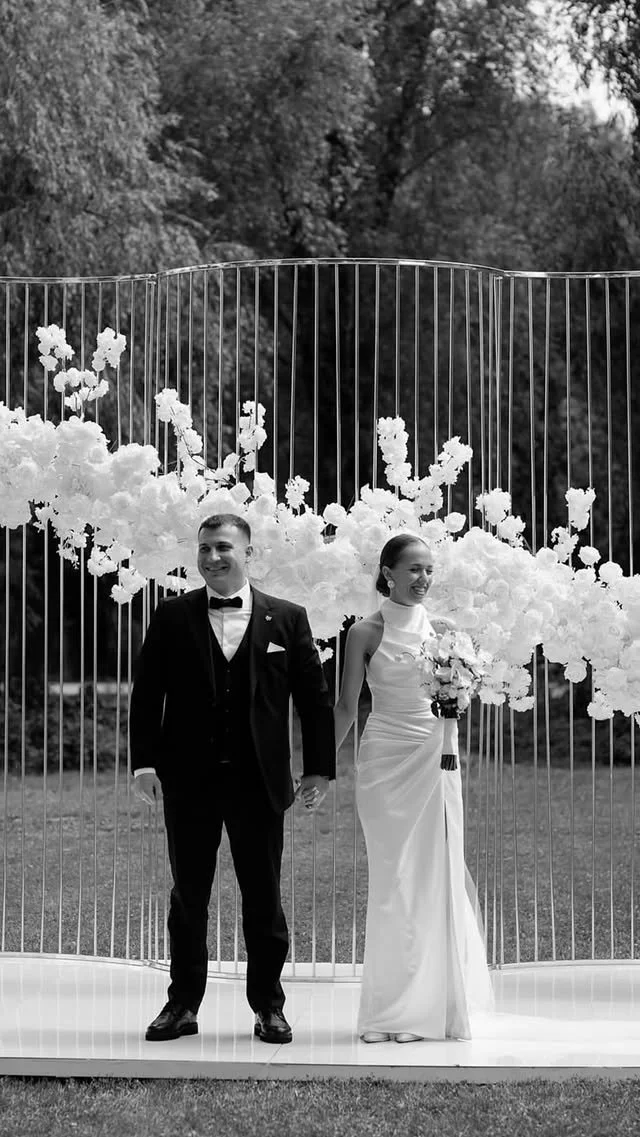 Black and white photo of a bride and groom holding hands at their outdoor wedding ceremony, standing in front of a floral backdrop with white flowers and a decorative fence, surrounded by trees.
