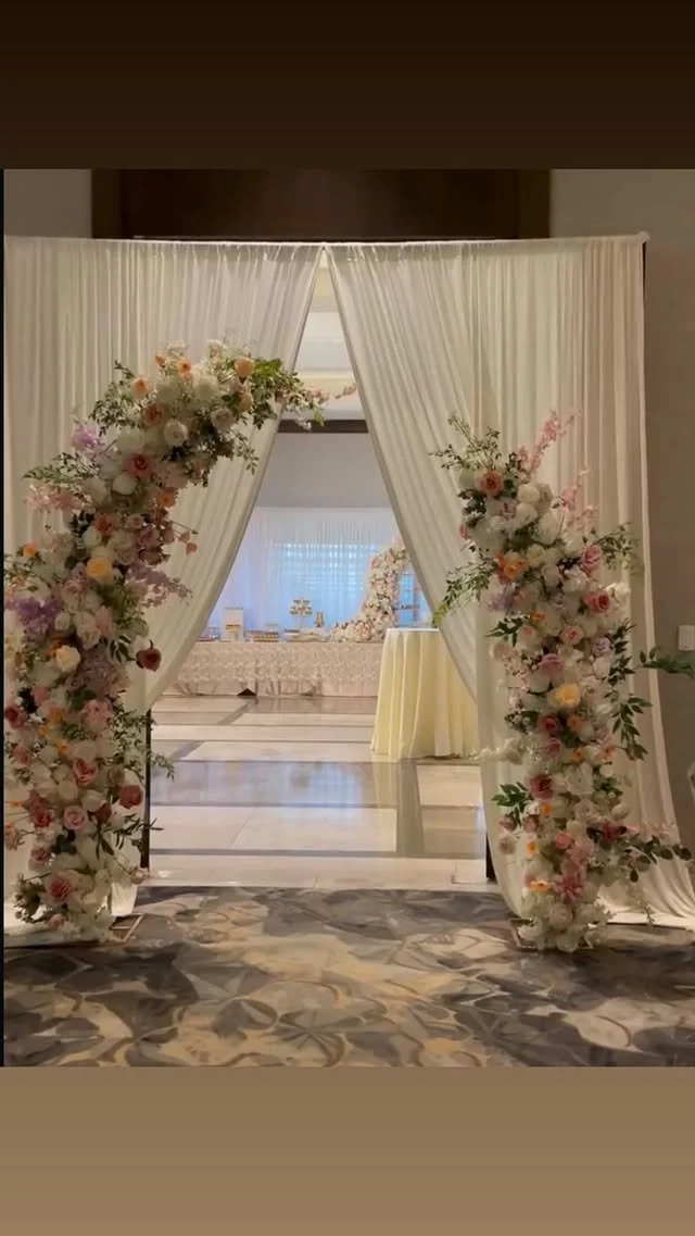 Elegant wedding arch decorated with pastel-colored flowers and white curtains in an indoor venue.