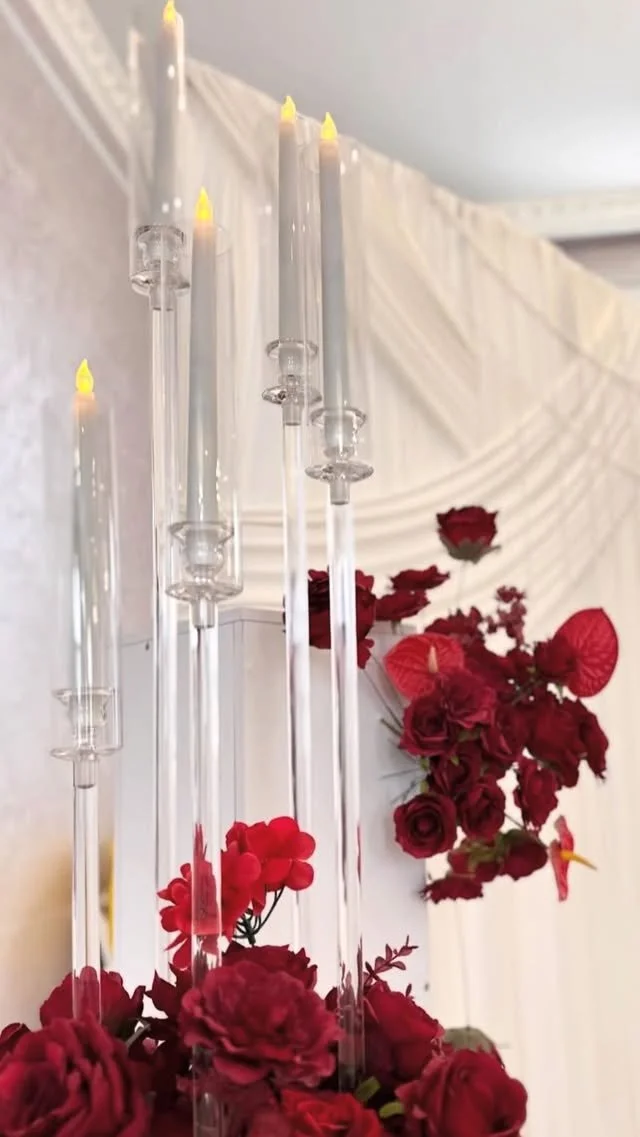 Several tall, lit candle sticks hanging vertically against a white draped background with red flowers underneath.