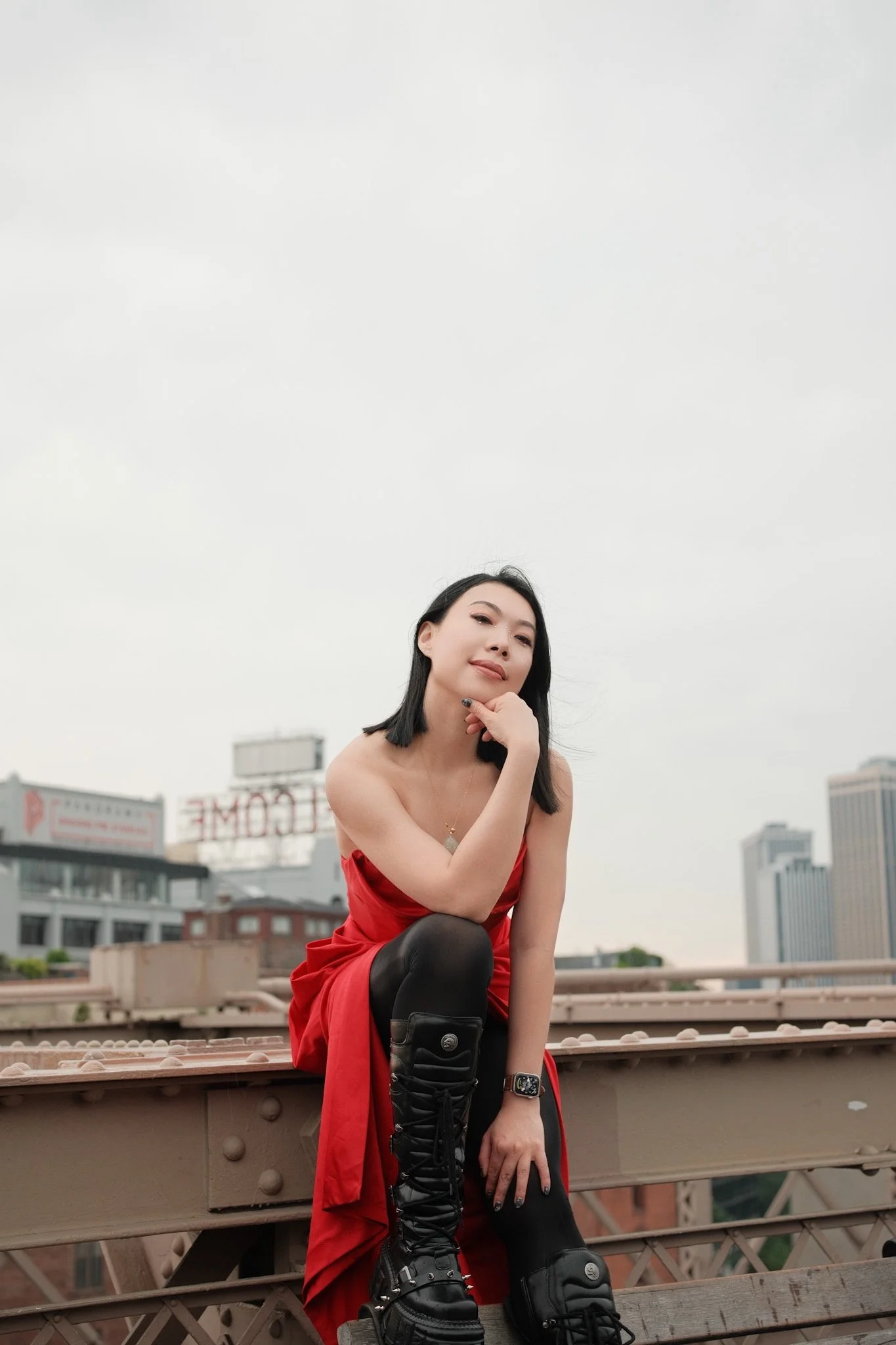 A woman in a red dress and black leggings with black lace-up boots is sitting on the edge of a bridge with an urban cityscape in the background. She has black hair and is looking into the camera, smiling slightly, with her left hand resting on her ch