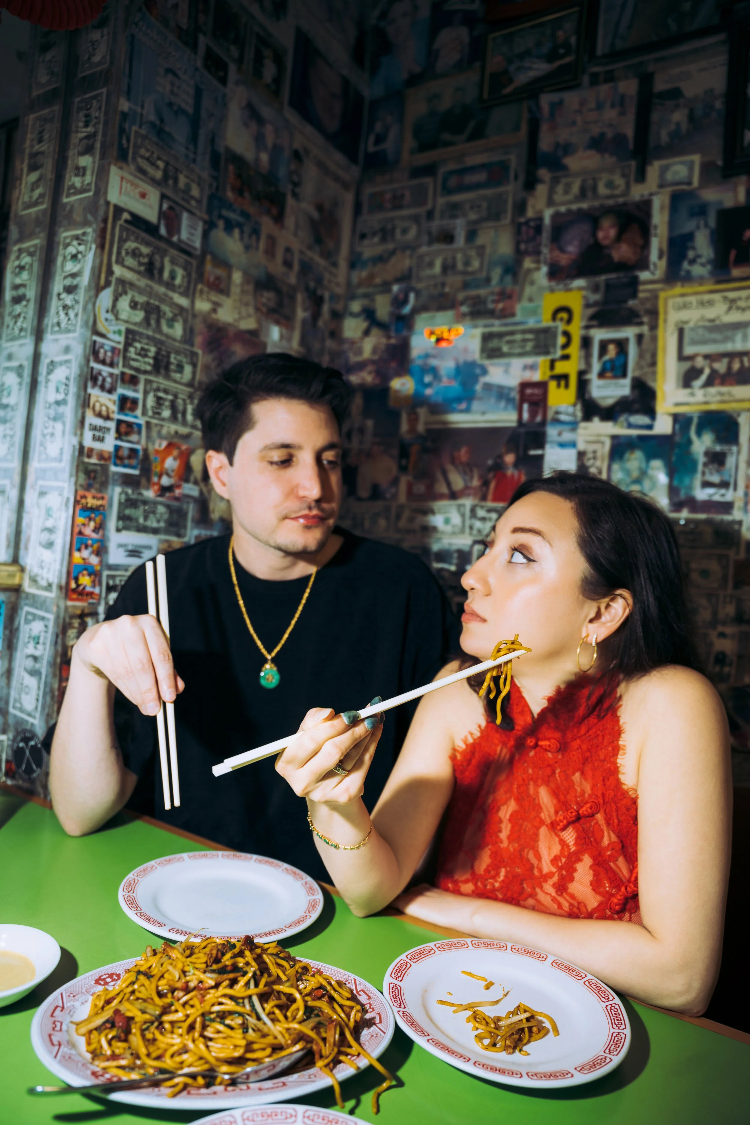 A man is feeding a woman chopsticks at a Chinese restaurant. The woman is dressed in a red lace dress, and plates of noodles are on a green table in front of them. The background features a wall covered with photos and memorabilia.