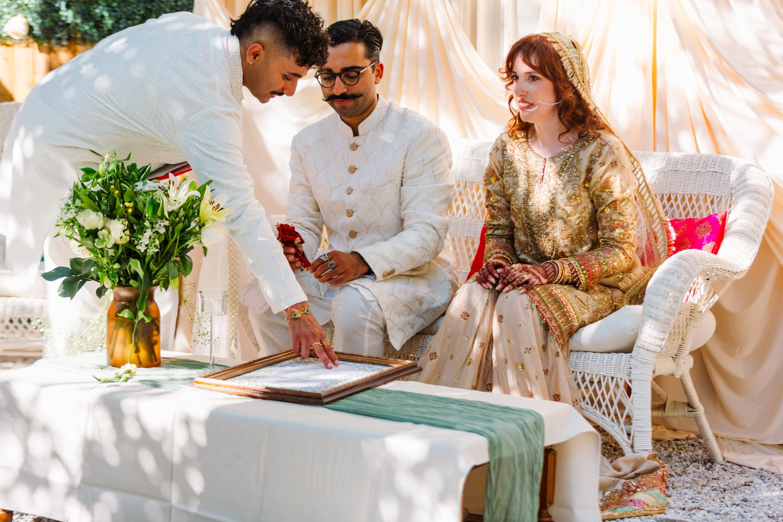 Three people dressed in traditional South Asian attire participating in a ceremony, with decorations in the background and a table with a floral arrangement in the foreground.