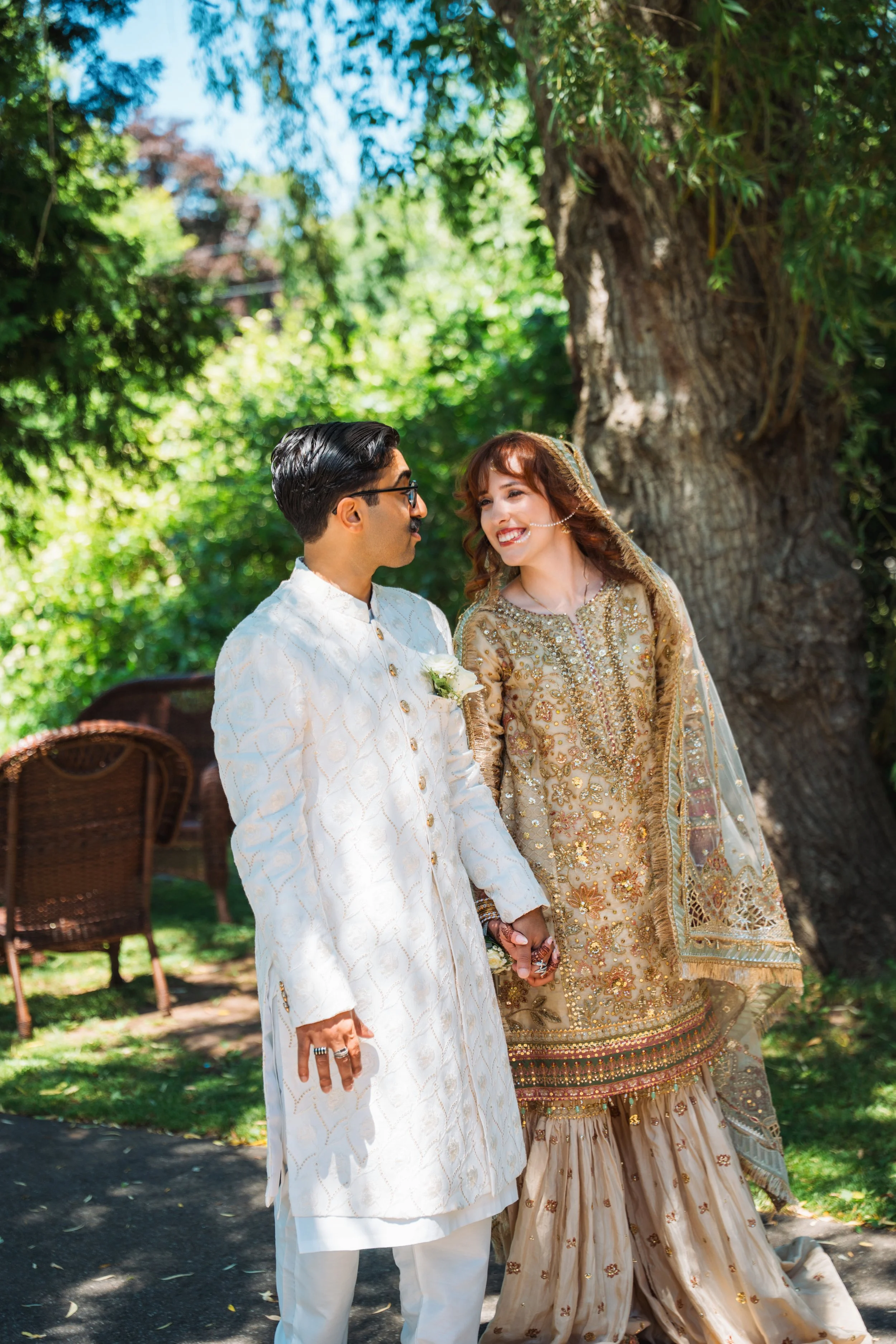 A couple dressed in traditional South Asian bridal attire holding hands outdoors under a large tree, with bright sunlight and green foliage.