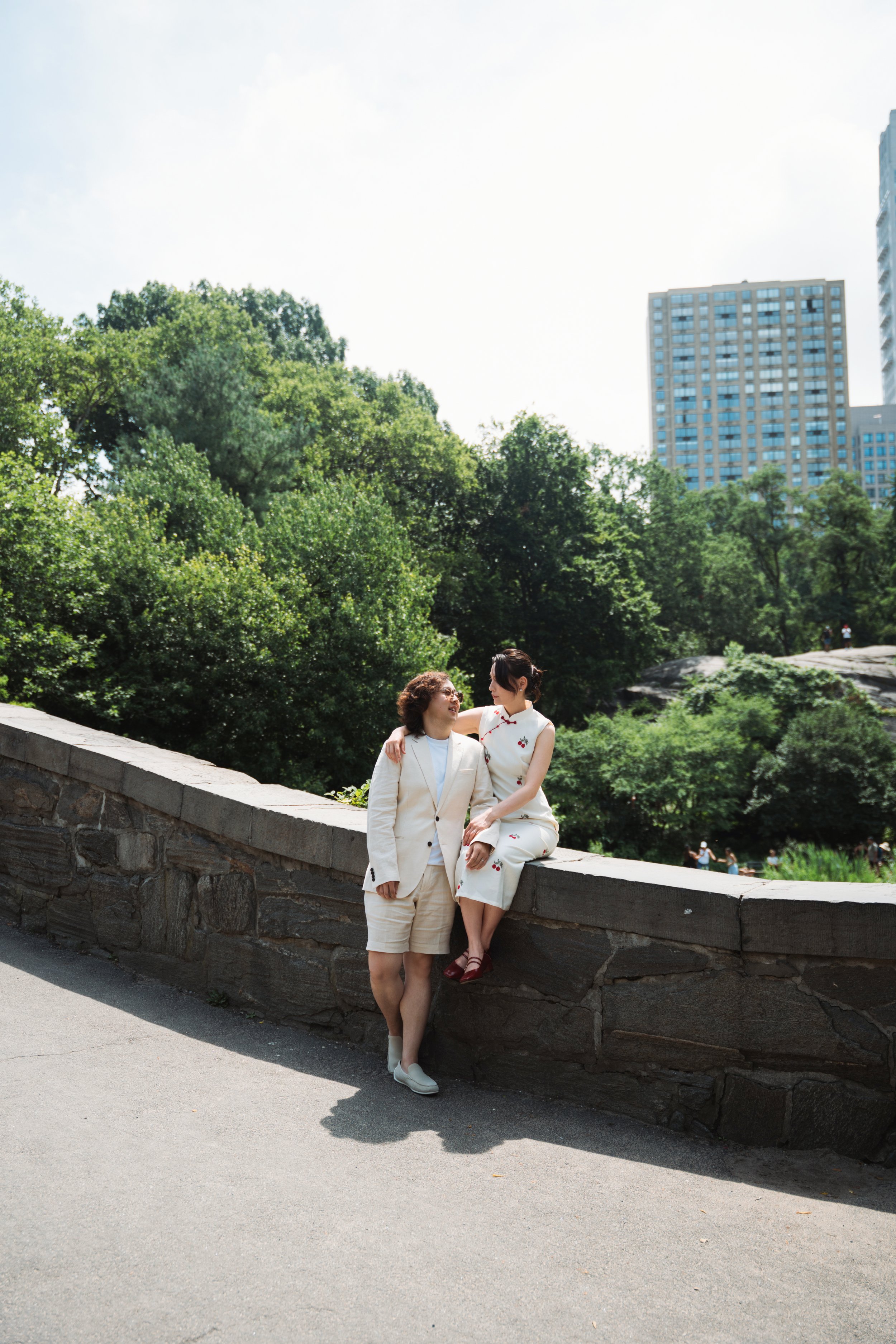 Two women dressed in formal attire are posing on a stone wall in a park with green trees and tall buildings in the background; one woman is standing and the other is sitting on the wall, and they are smiling at each other.