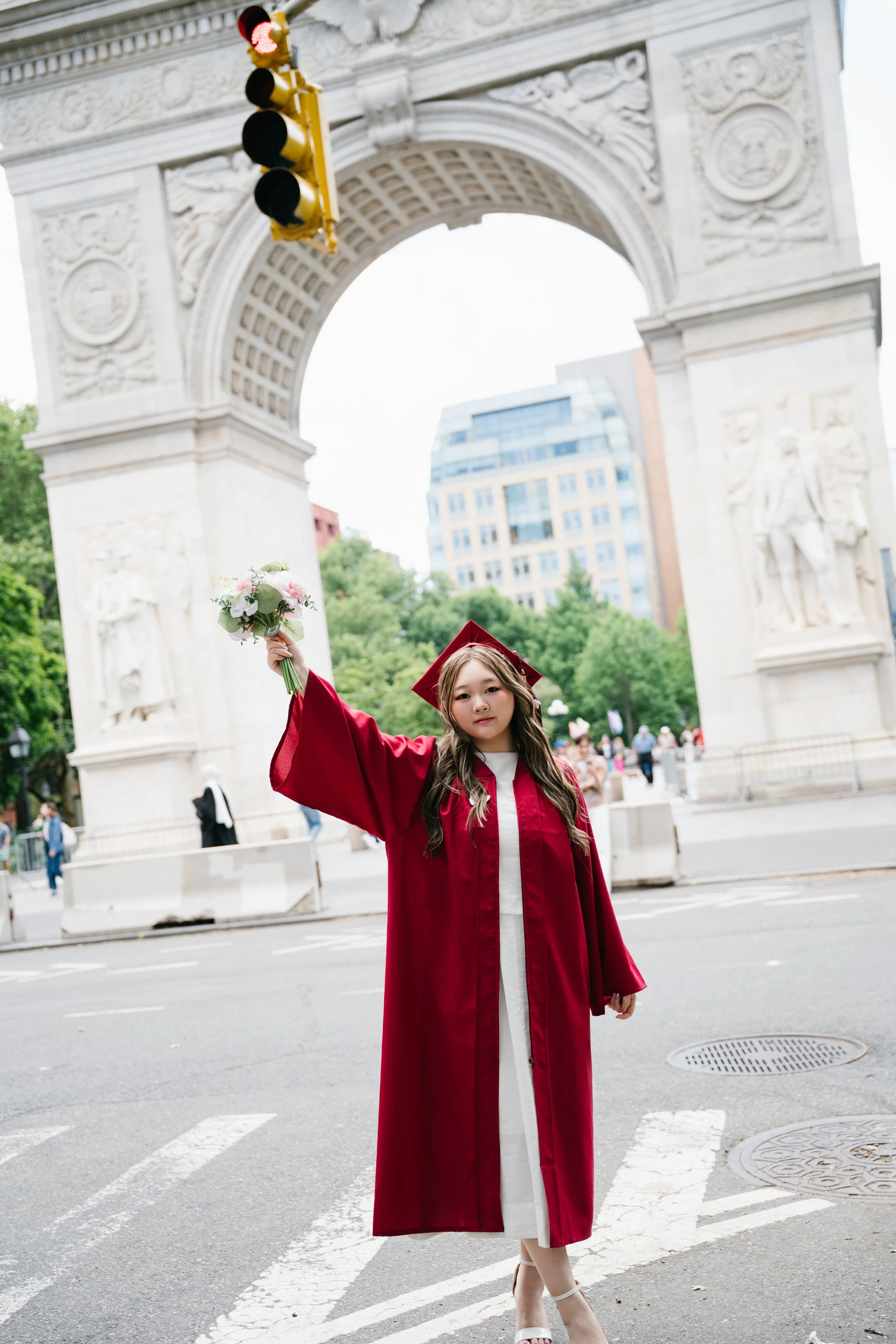 A young woman in a red graduation gown and cap holding a bouquet of flowers, standing on a city street in front of an ornate stone archway.