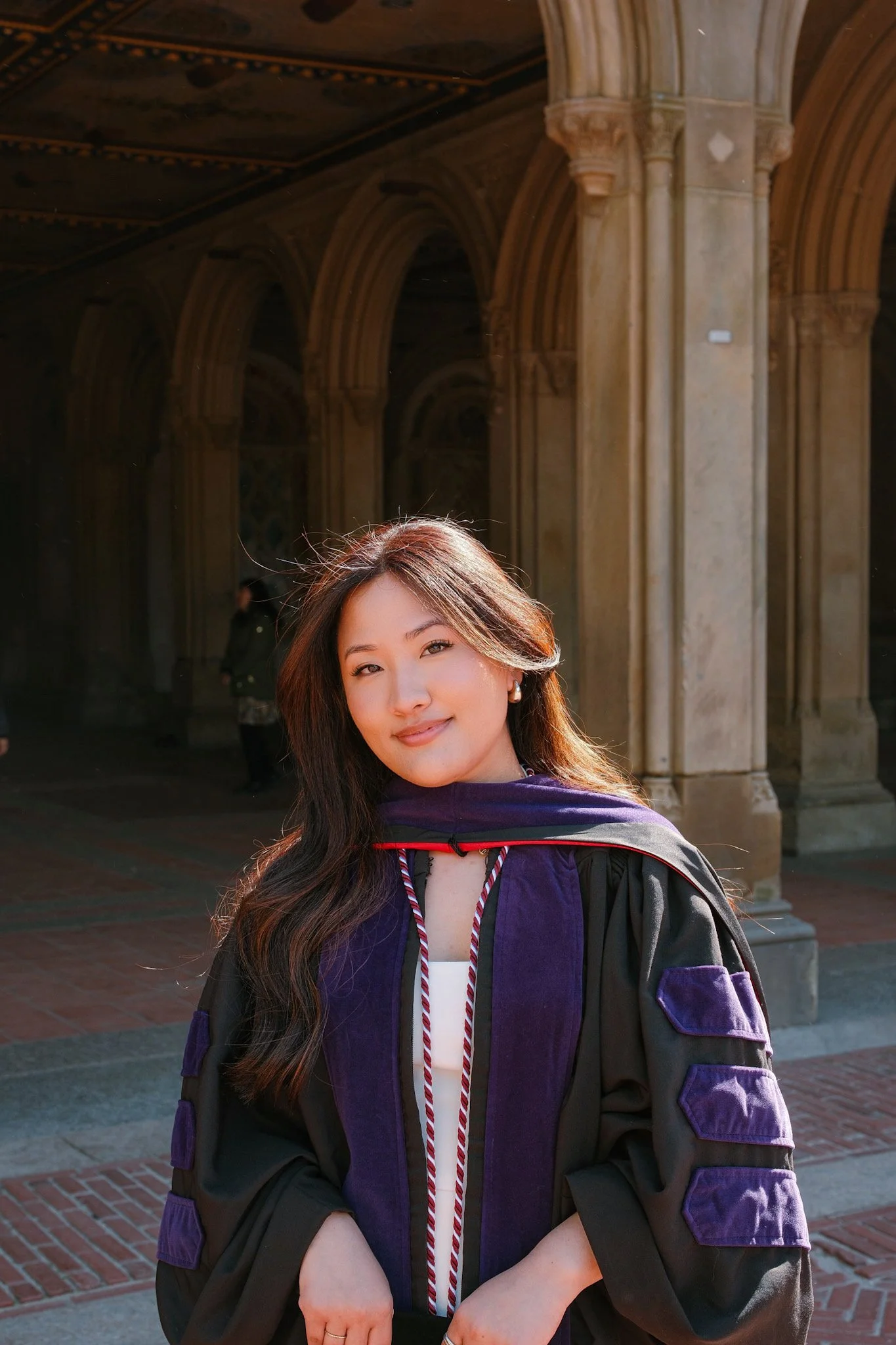 A woman with long, wavy brown hair and light makeup is smiling gently at the camera. She is wearing a black graduation gown with purple accents and a white shirt underneath. The background shows stone columns and arches, indicating an academic or his