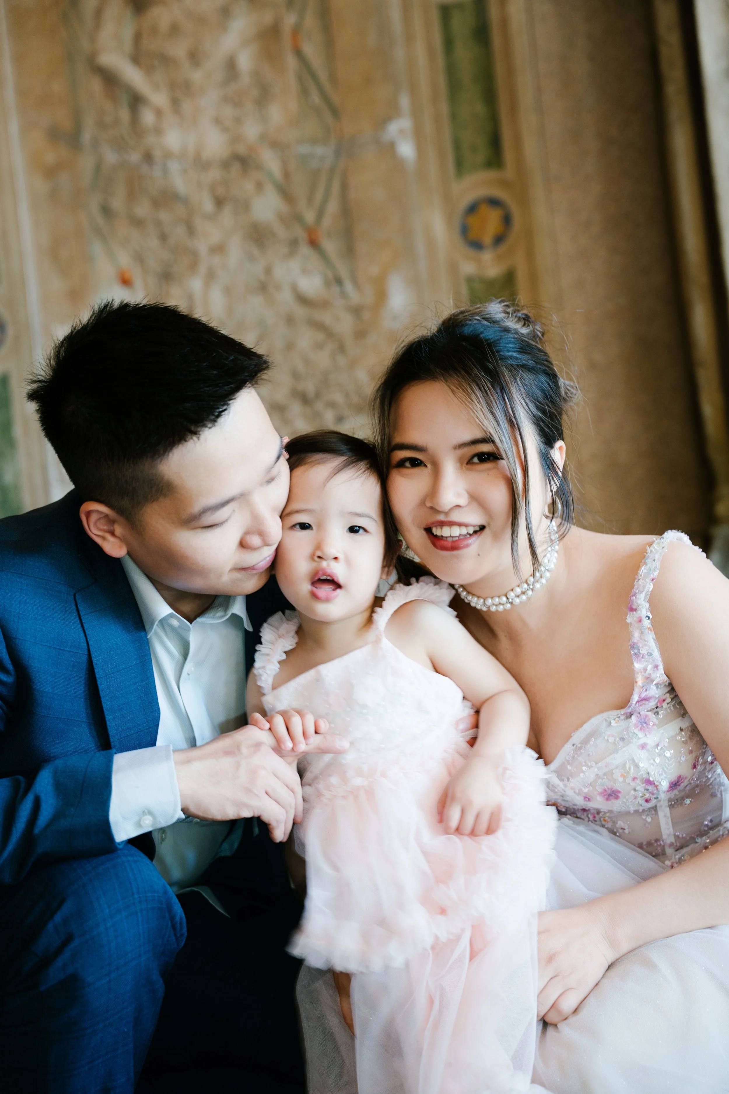 A happy family portrait featuring a man, woman, and young girl dressed in formal attire, smiling and sitting close together in an ornately decorated room.