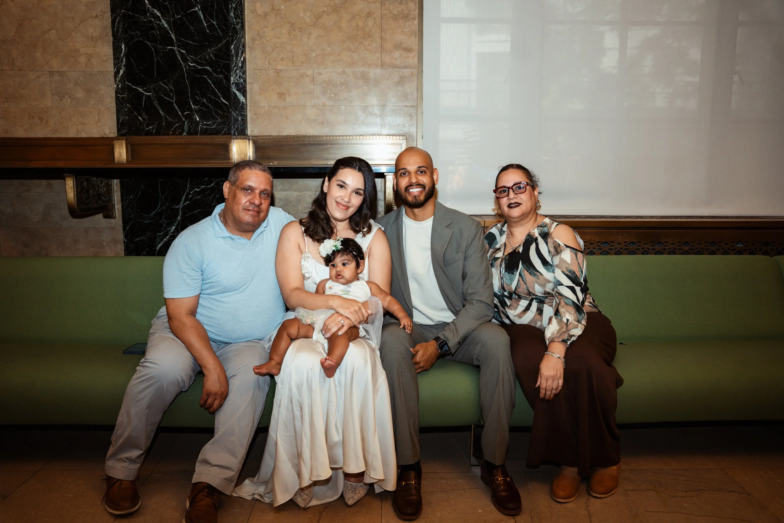 A diverse group of five adults and one child sitting on a green bench in an indoor setting with marble walls and a glass window in the background.
