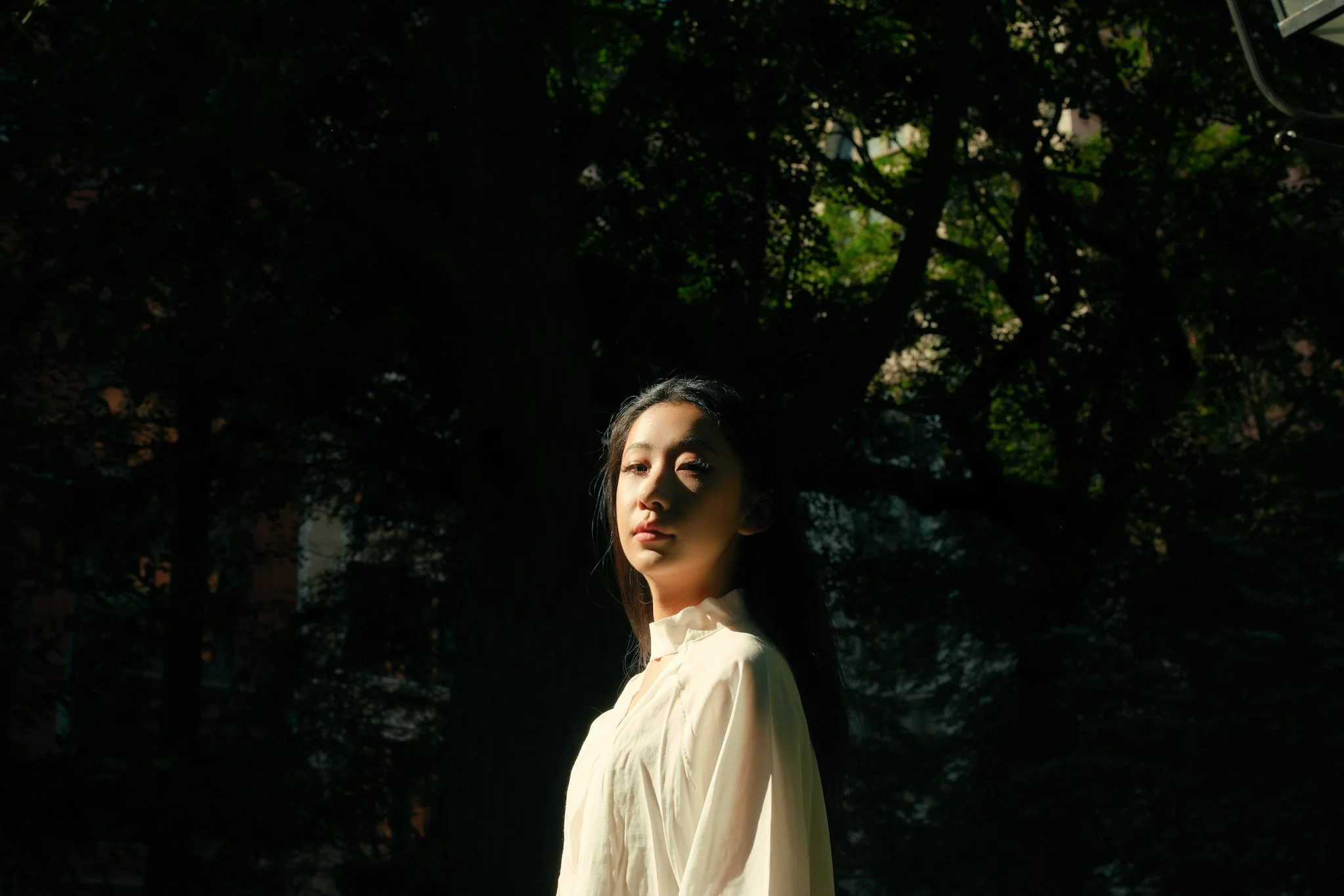 Portrait of a young woman with dark hair and fair skin, standing outdoors under a large tree with dense foliage, wearing a cream-colored top, with sunlight highlighting her face.