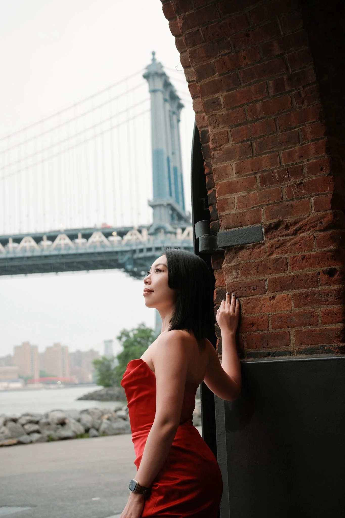 A woman with dark hair wearing a red dress and a smartwatch on her left wrist, leaning against a brick wall near the waterfront with a bridge in the background.