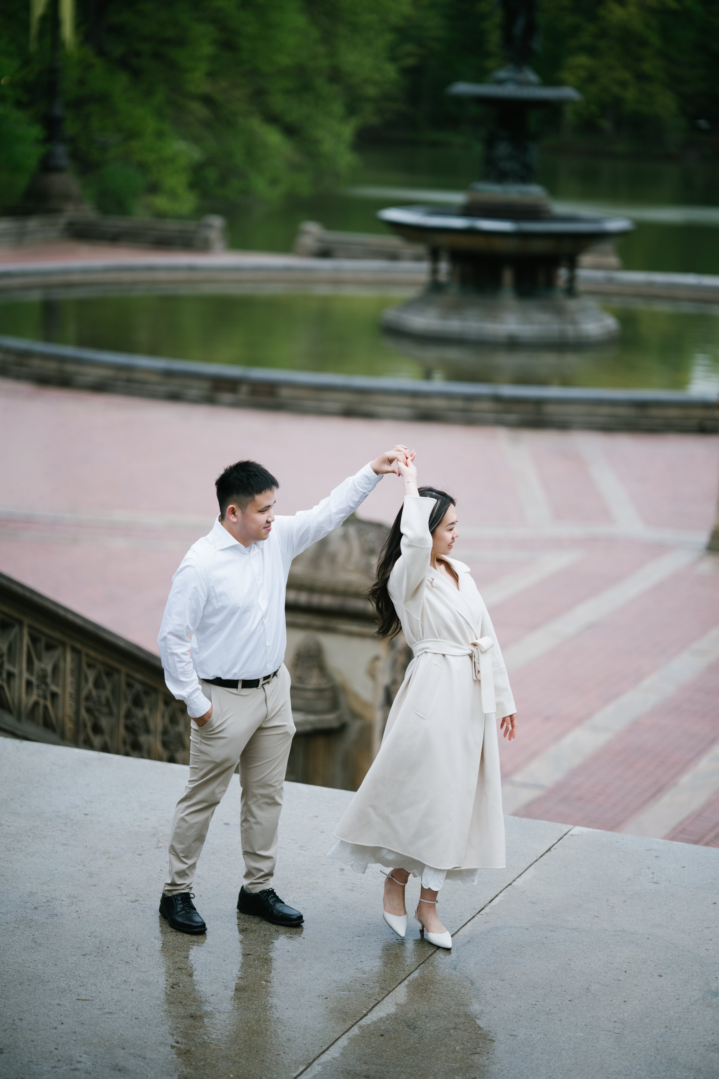 A young couple dancing outdoors near a fountain in a park, with the man holding the woman's hand as she walks forward in a white dress and high heels, the man dressed in beige pants and a white shirt, on a cloudy day.