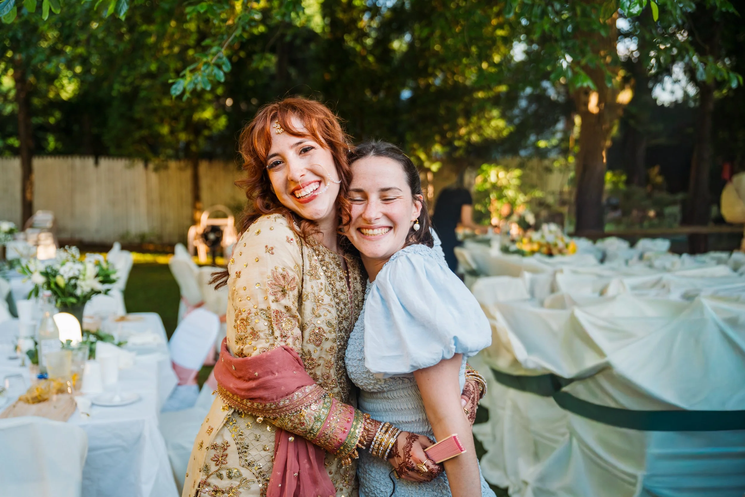 Two women embracing and smiling at the camera during an outdoor wedding reception, with decorated tables and a wooden fence in the background.