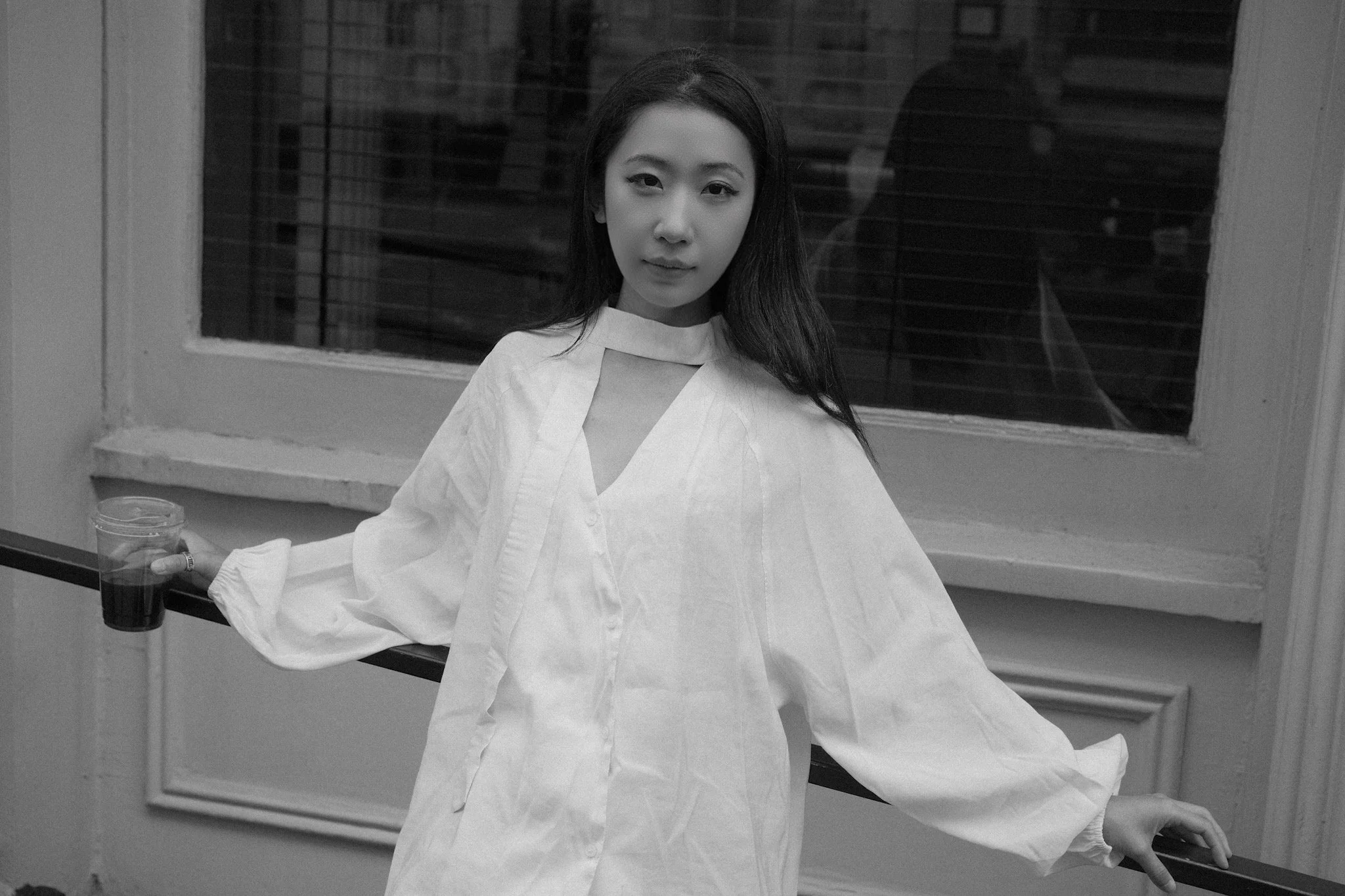 A young woman with long dark hair and light skin wearing a white garment, standing in front of a window with blinds inside a room. She is holding a dark-colored drink with a straw in her right hand. The scene appears to be at night outside.