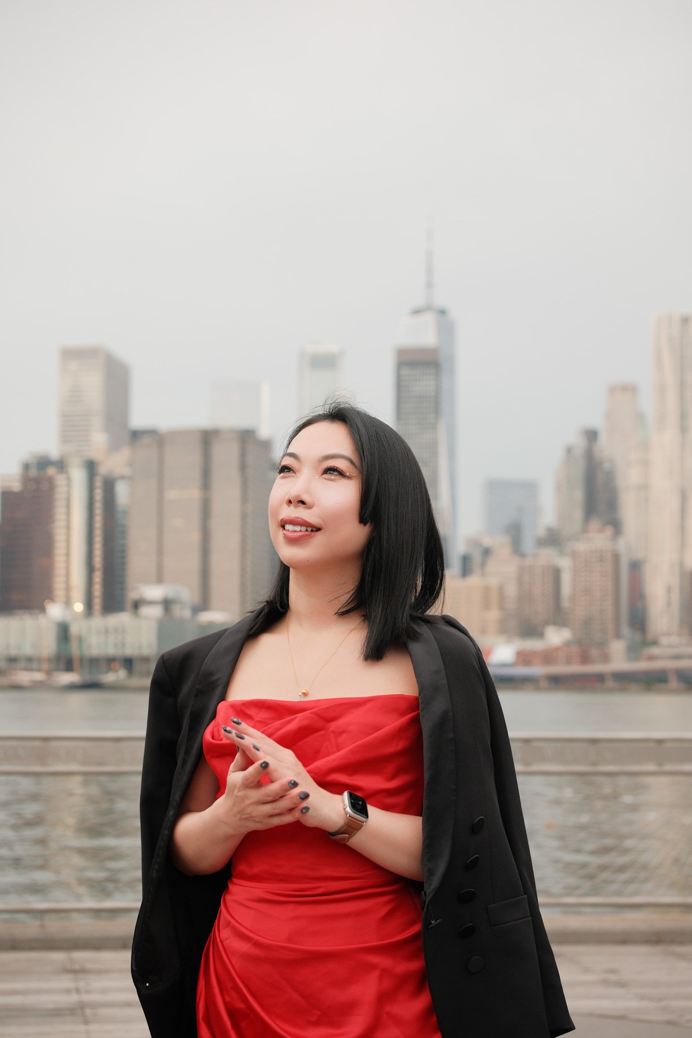 A woman with black shoulder-length hair and light skin is standing outdoors in front of a city skyline, including tall buildings like the One World Trade Center, with water and a railing behind her. She is smiling softly, looking slightly upwards, an