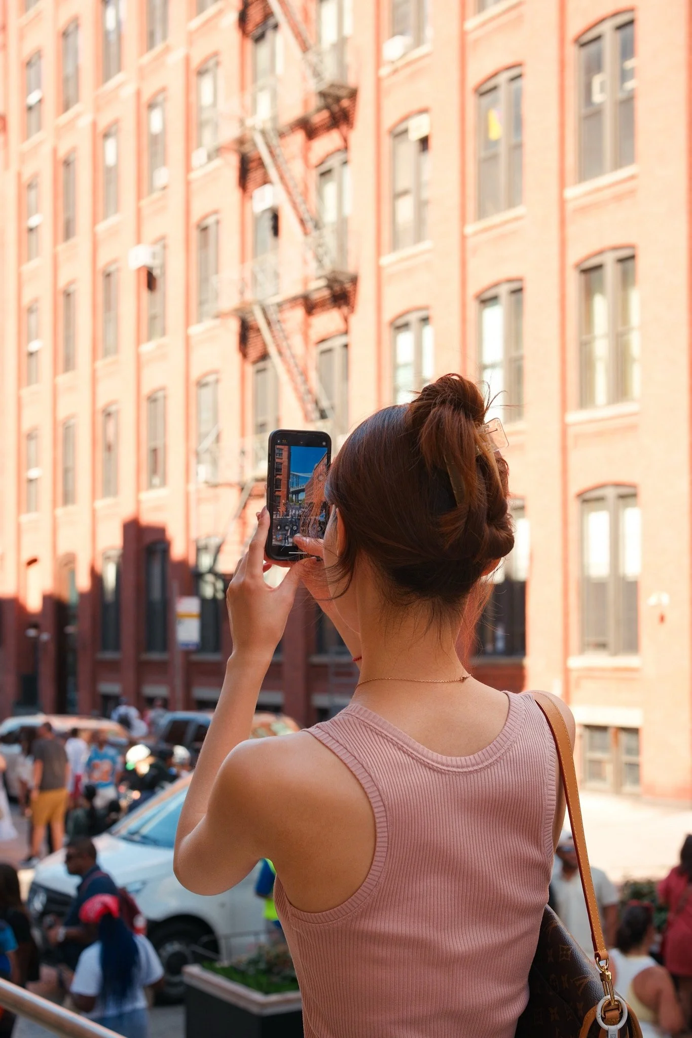 A woman with brown hair styled in a bun is taking a photo with her smartphone in a busy city street. She is wearing a pink sleeveless top and carrying a Louis Vuitton handbag. The background shows a crowd of people and a row of multi-story brick buil