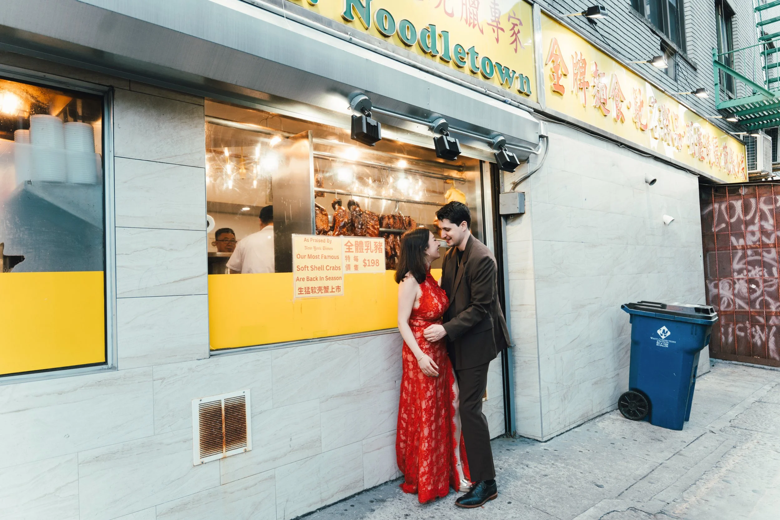 A couple dressed in formal attire, a woman in a red lace dress and a man in a dark suit, standing close and smiling outside a restaurant with a yellow signboard that says "NoodelTown". Inside the restaurant, roasted meats are visible through the wind