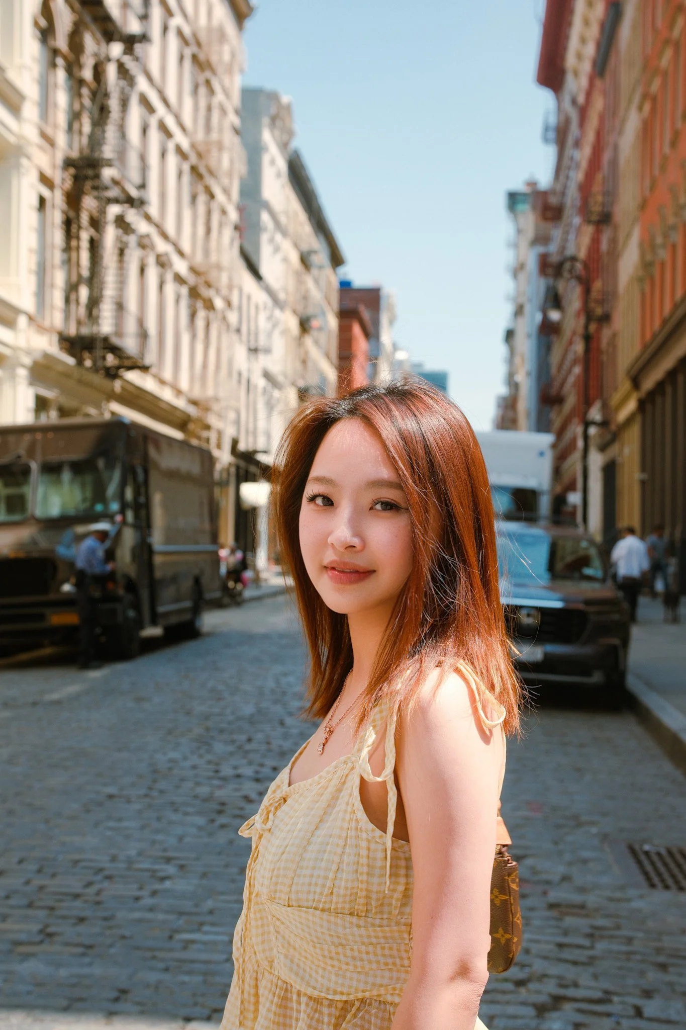 Young woman with reddish-brown hair standing on a cobblestone city street with buildings on either side, some cars and people in the background, smiling slightly.