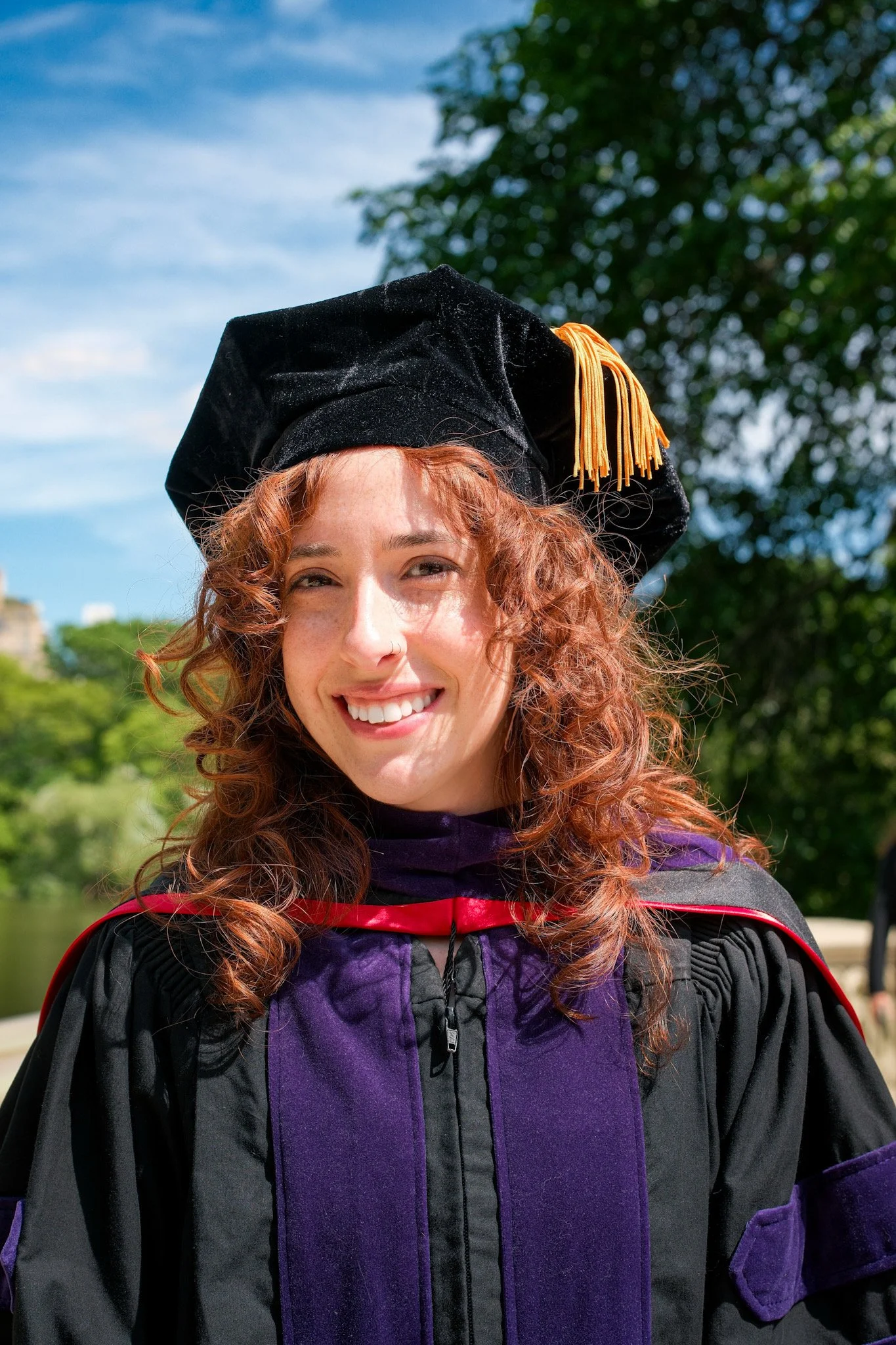 A young woman with red curly hair wearing a graduation cap and gown outdoors with a background of blue sky, trees, and greenery.