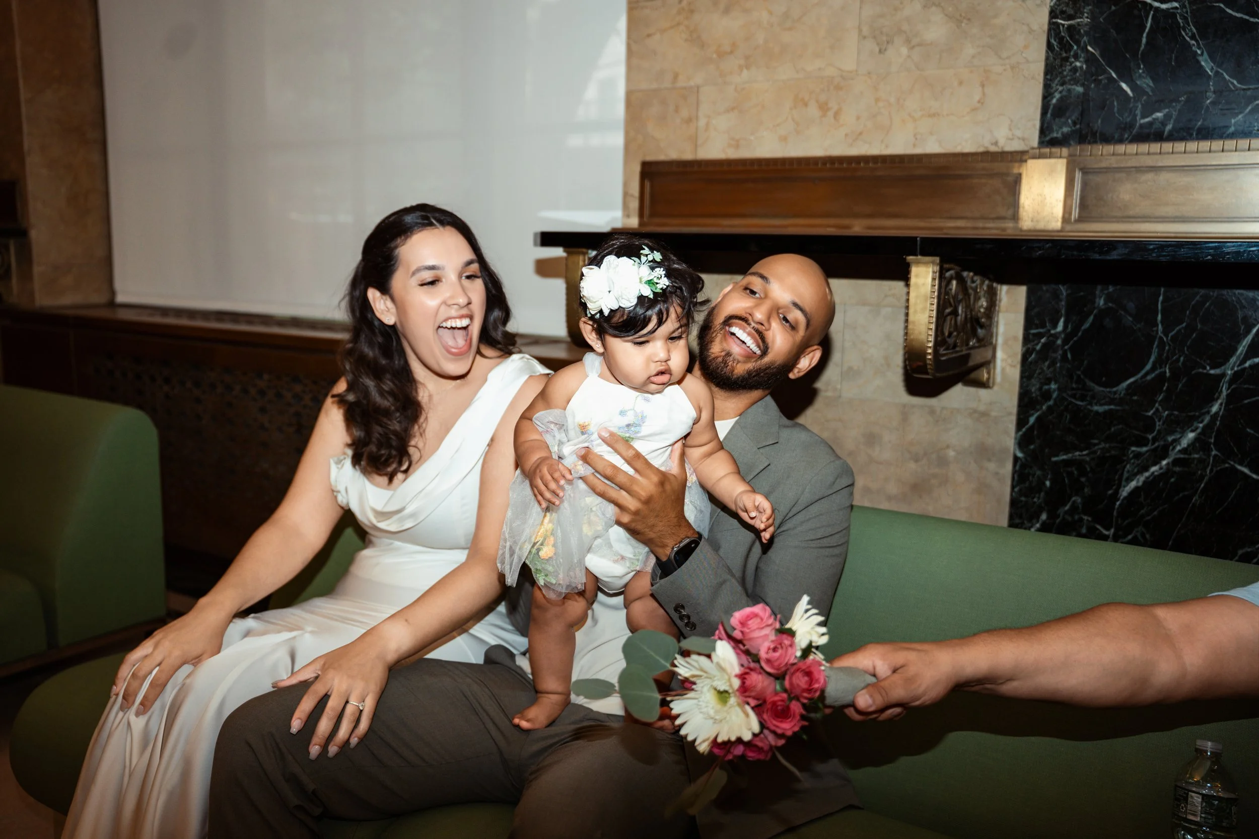 A happy family of three, including a woman, a man, and a young girl, sitting together on a green sofa in a well-decorated room, holding a bouquet of flowers, with the woman laughing and the man smiling.