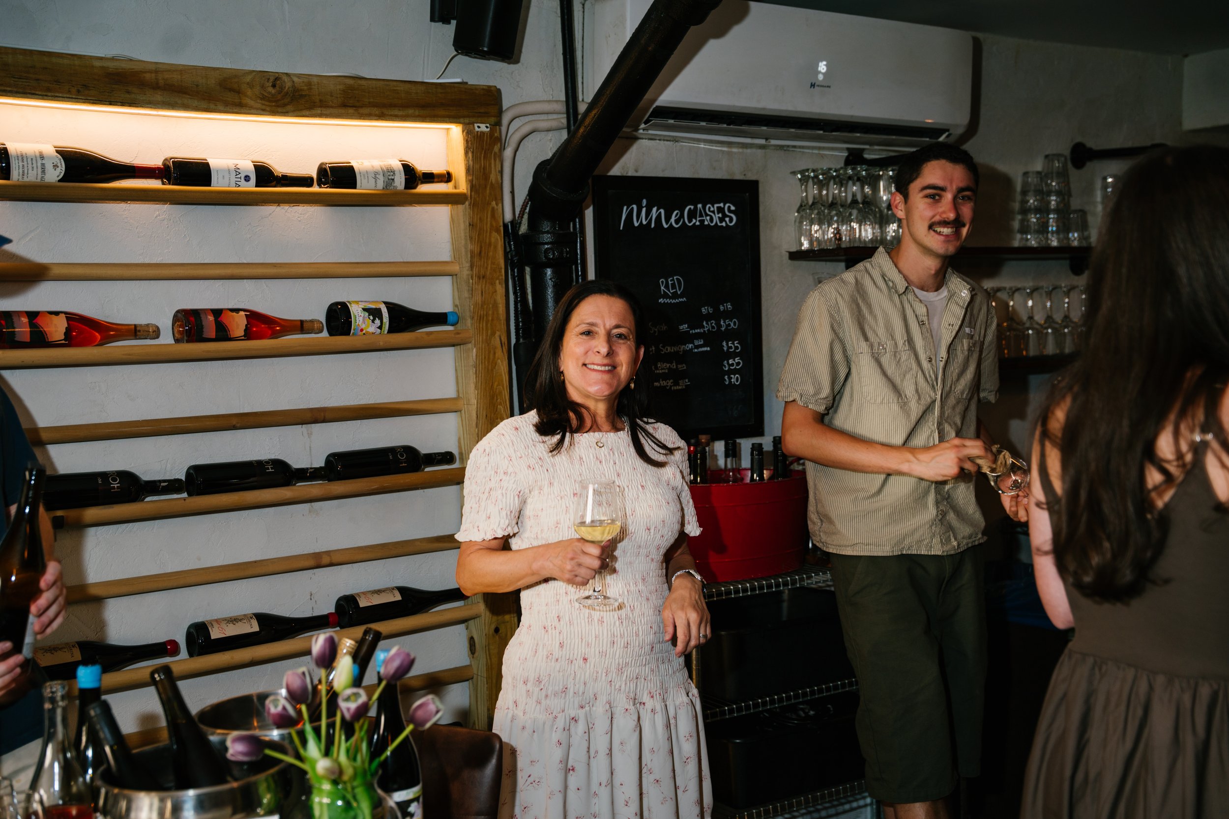 Women and young man at a bar or restaurant, woman holding a glass of white wine, smiling, wine bottles on shelves behind, chalkboard menu listing wines, glasses on shelves, decorative tulips in a container, casual setting.