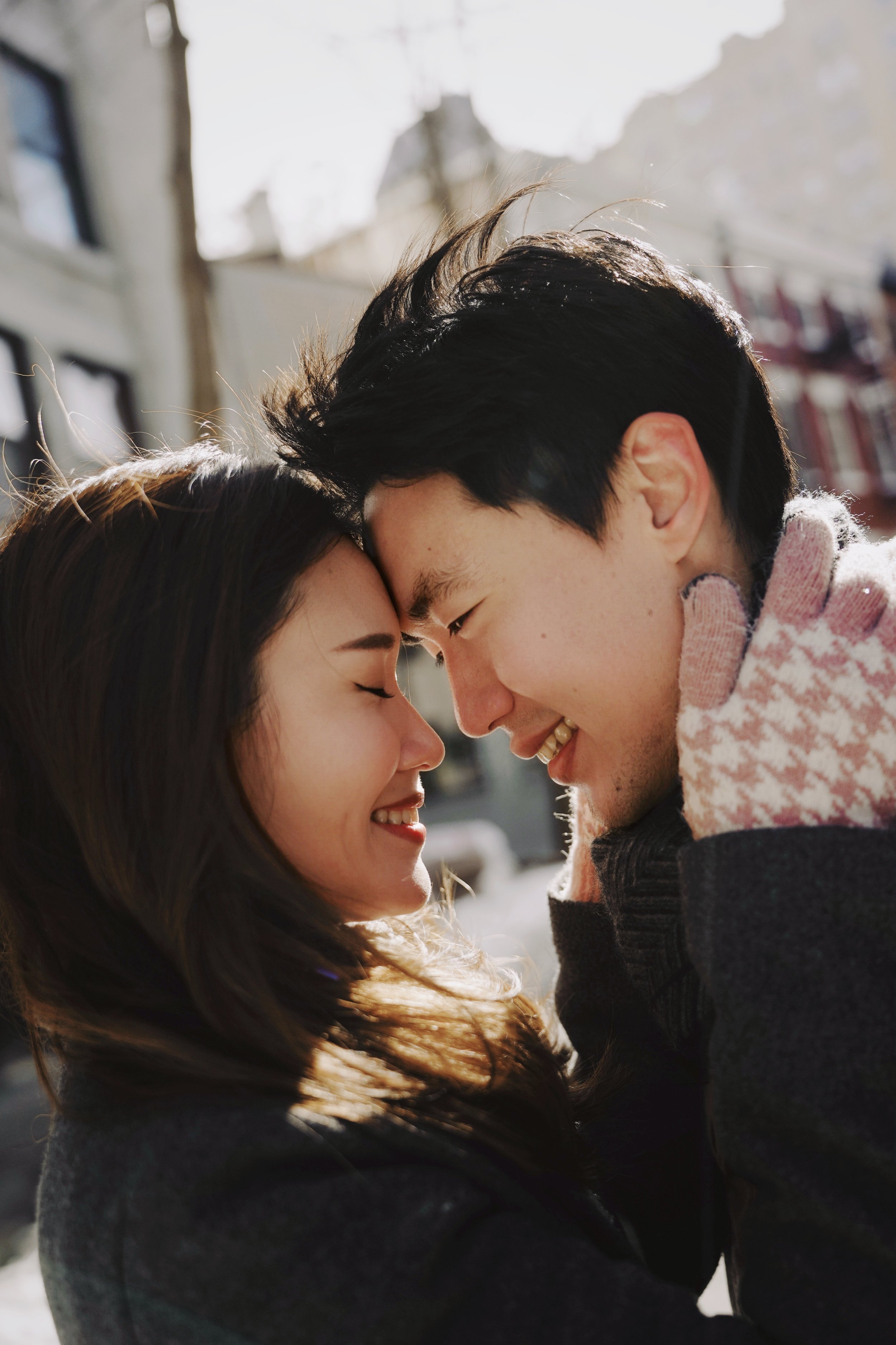 A young couple with their foreheads touching, smiling with closed eyes in an outdoor setting during daylight, with buildings in the background.