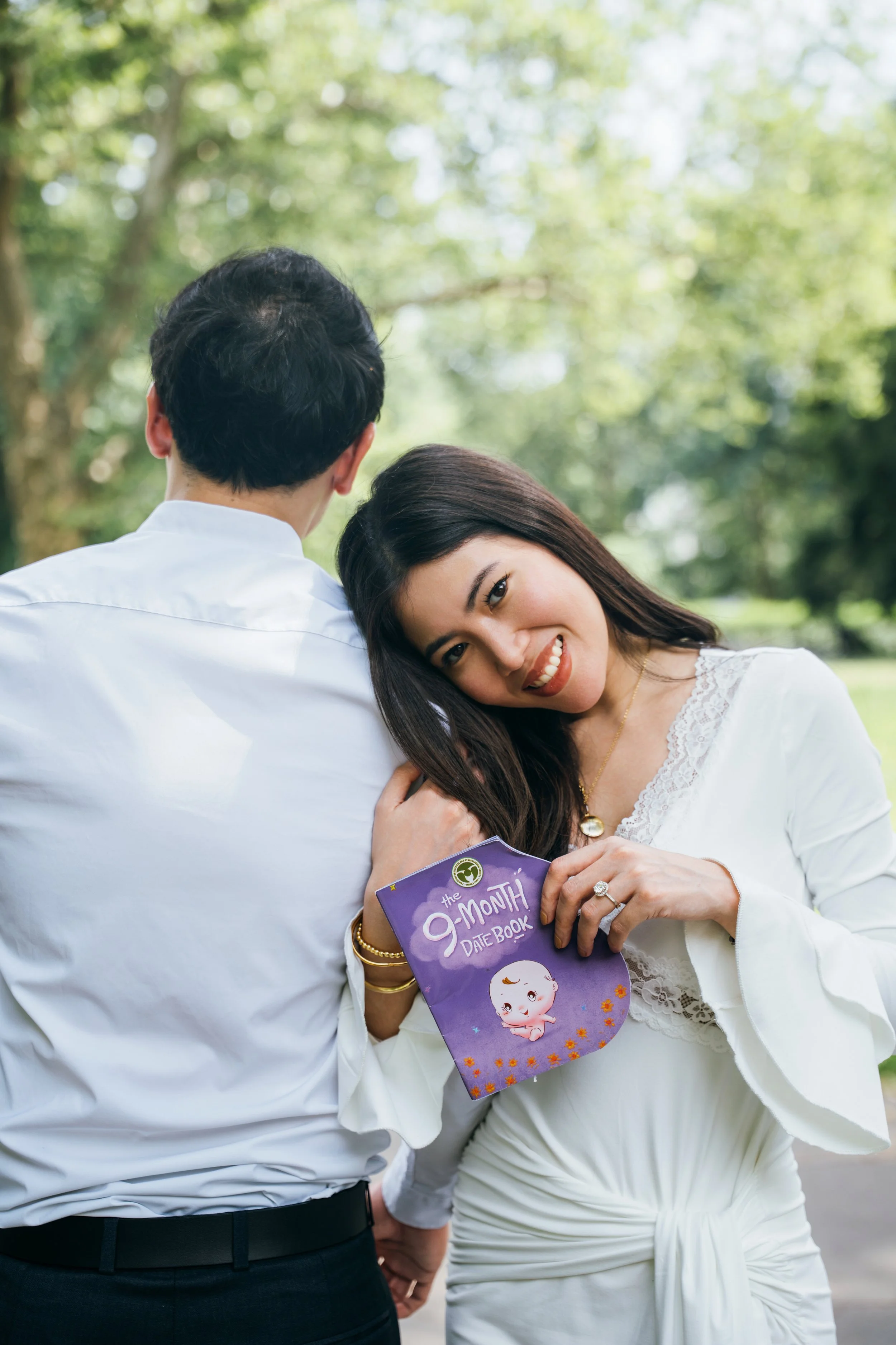 A woman smiling and resting her head on a man's shoulder outdoors, holding a purple 9-month date book with a cartoon baby on the cover.