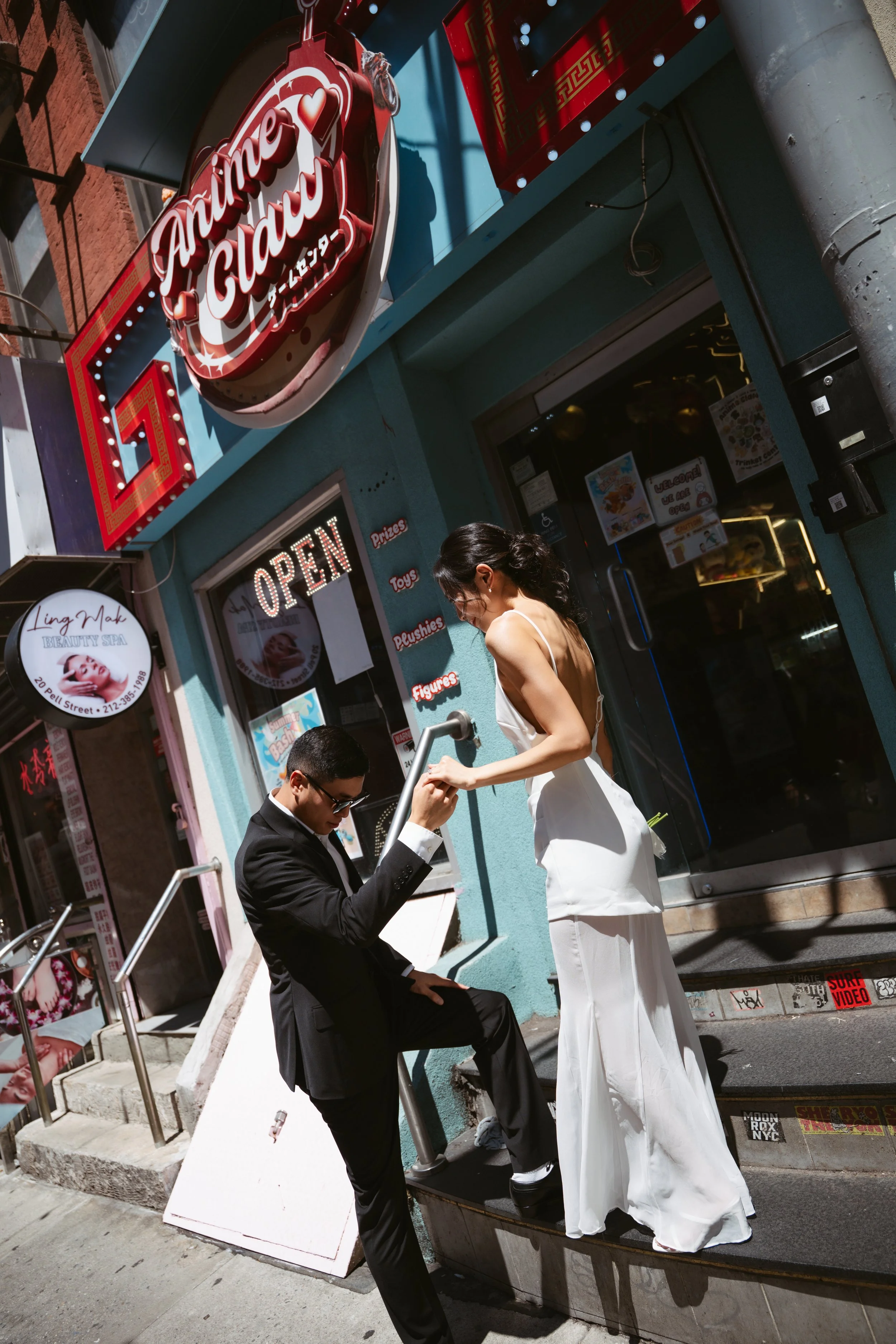 A woman in a white dress and a man in a black suit are outside a colorful storefront with a red sign that reads 'Anime Glam.' The woman is standing on the steps, while the man is sitting on the steps, looking at something he is holding. The storefron