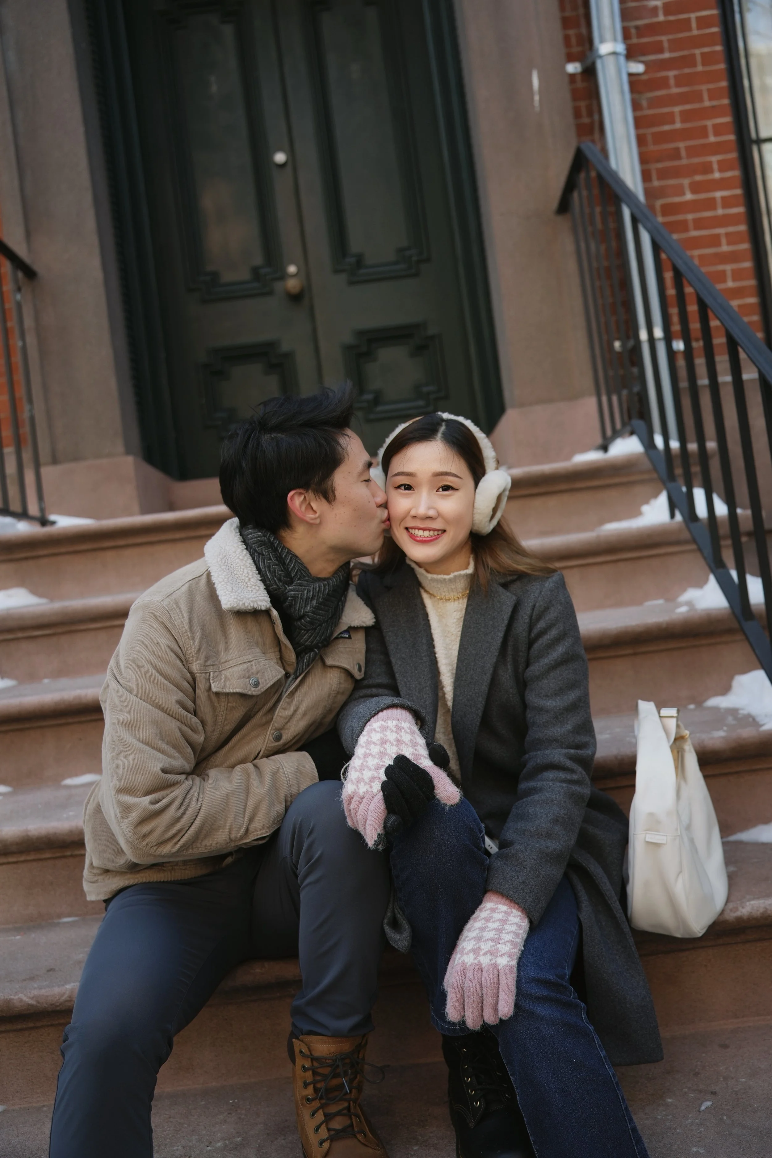 A young man kisses a young woman on the cheek while sitting on brownstone stairs outside a brick building in winter. The woman is smiling and wearing earmuffs, while the man wears a scarf and boots.