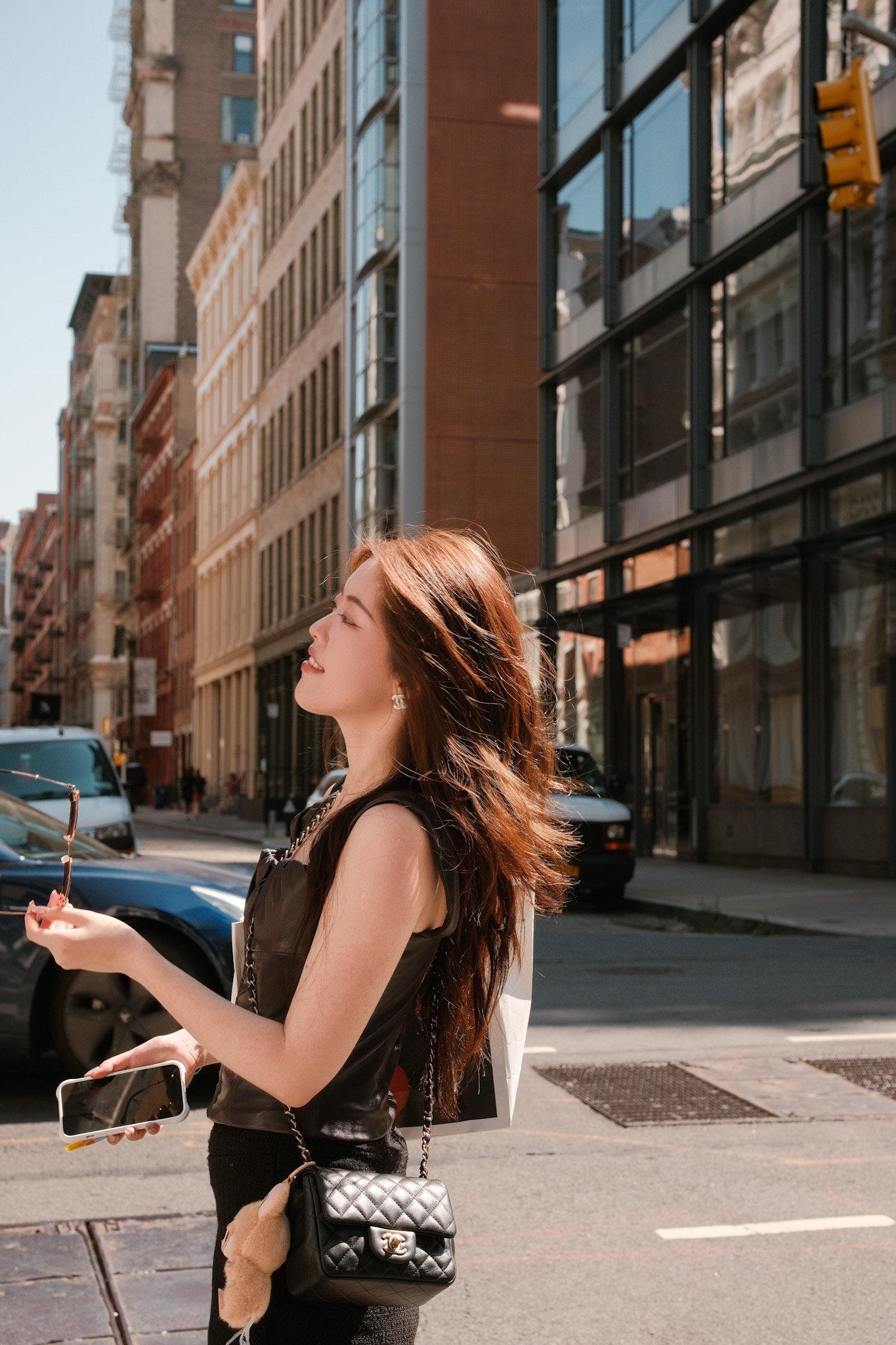 A young woman with long auburn hair, wearing a black sleeveless top, holding a smartphone in one hand and sunglasses in her other, standing on a city street with buildings and cars in the background.