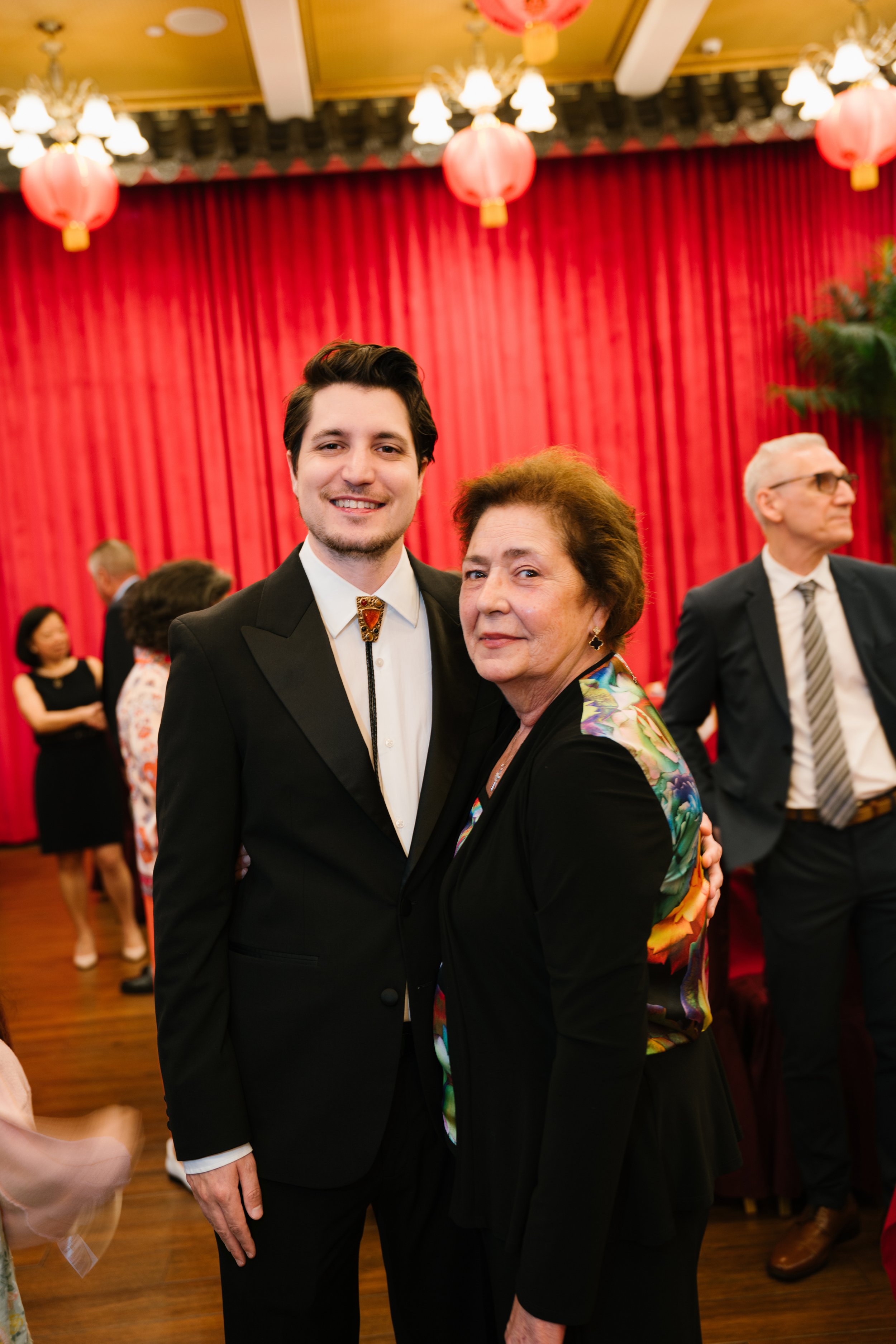 Young man in a tuxedo smiling and an older woman embracing at a celebration with red curtains and hanging lanterns in the background.