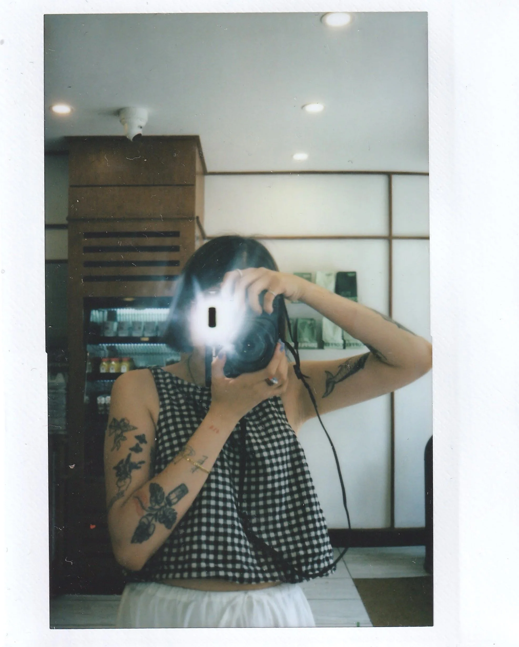 Person taking a mirror selfie, wearing a gingham top, with tattoos on their arm and holding a phone with a camera flash, in a room with white walls and ceiling lights.