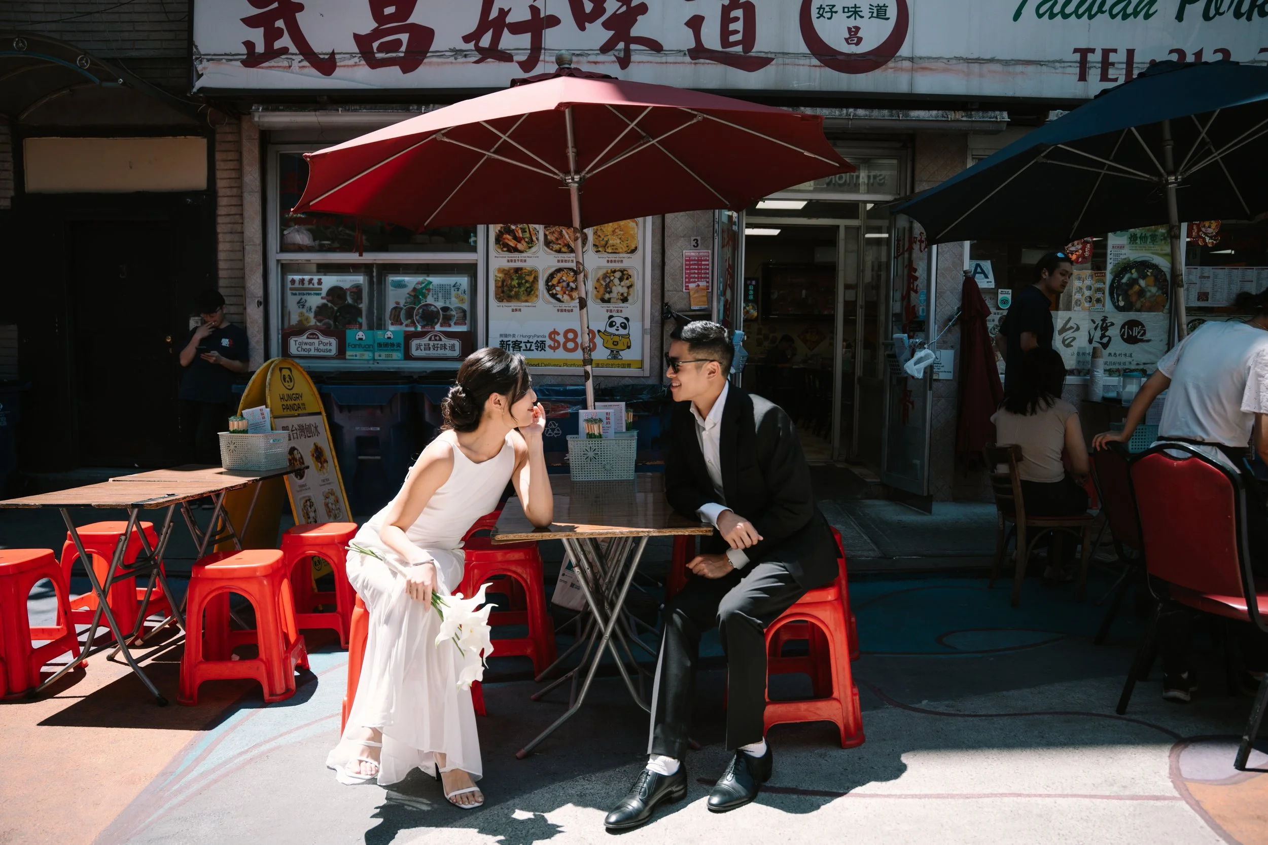 A couple with light skin and dark hair, dressed in white and black respectively, sitting at a red outdoor table at a Taiwanese restaurant, with red and blue umbrellas overhead. The woman is wearing a white dress and holding white flowers, and the man